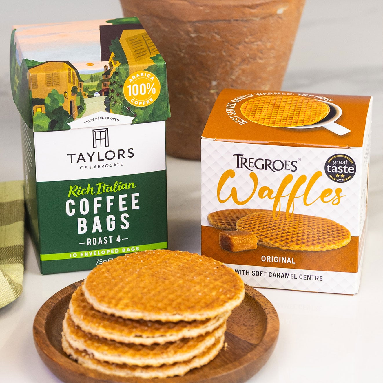 Taylors of Harrogate Rich Italian coffee bags paired with Tregroes Original stroopwafels stacked on a wooden plate beside a potted rosemary plant.