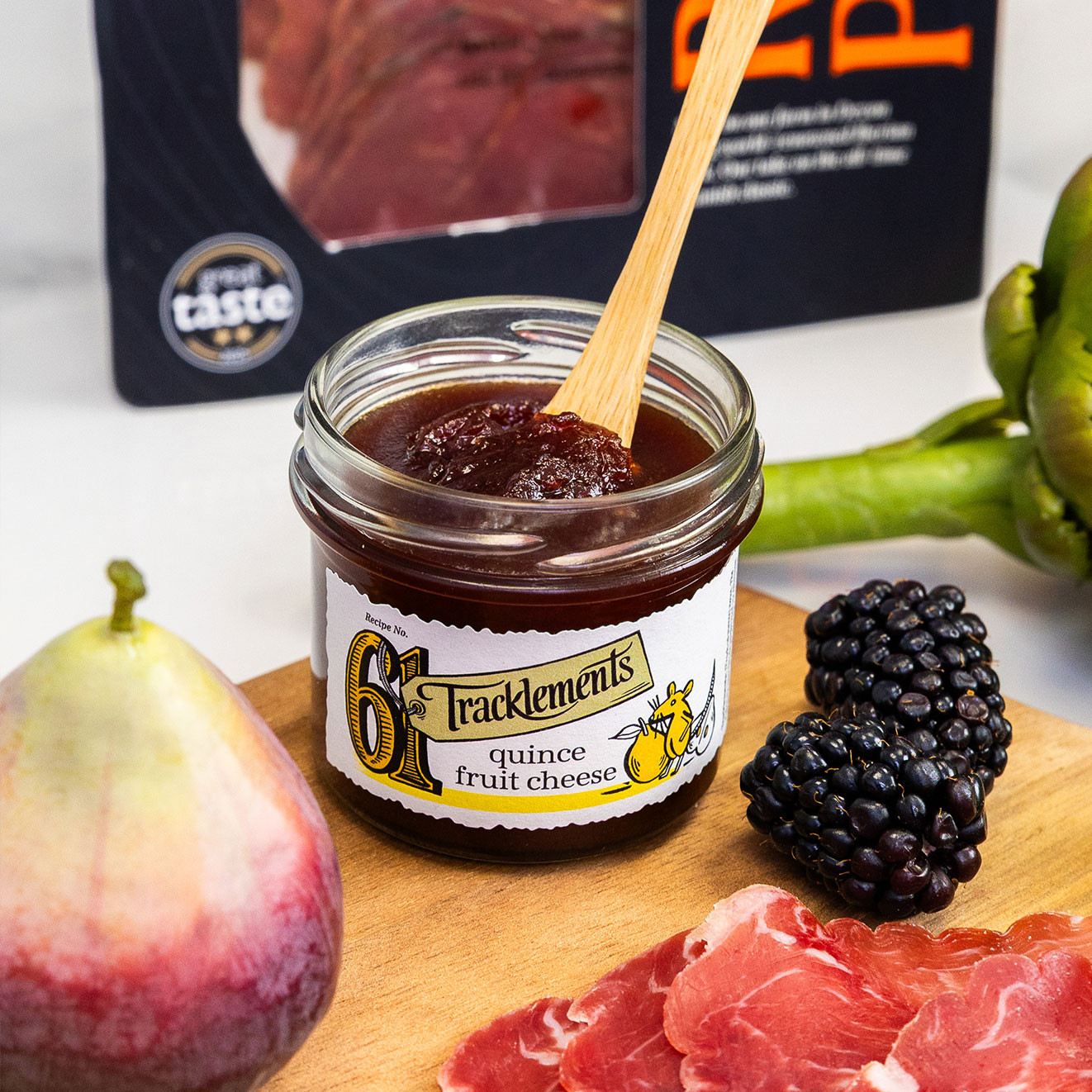 Tracklements quince fruit cheese in an open jar with a serving spoon, surrounded by blackberries, figs and slices of cured ham.