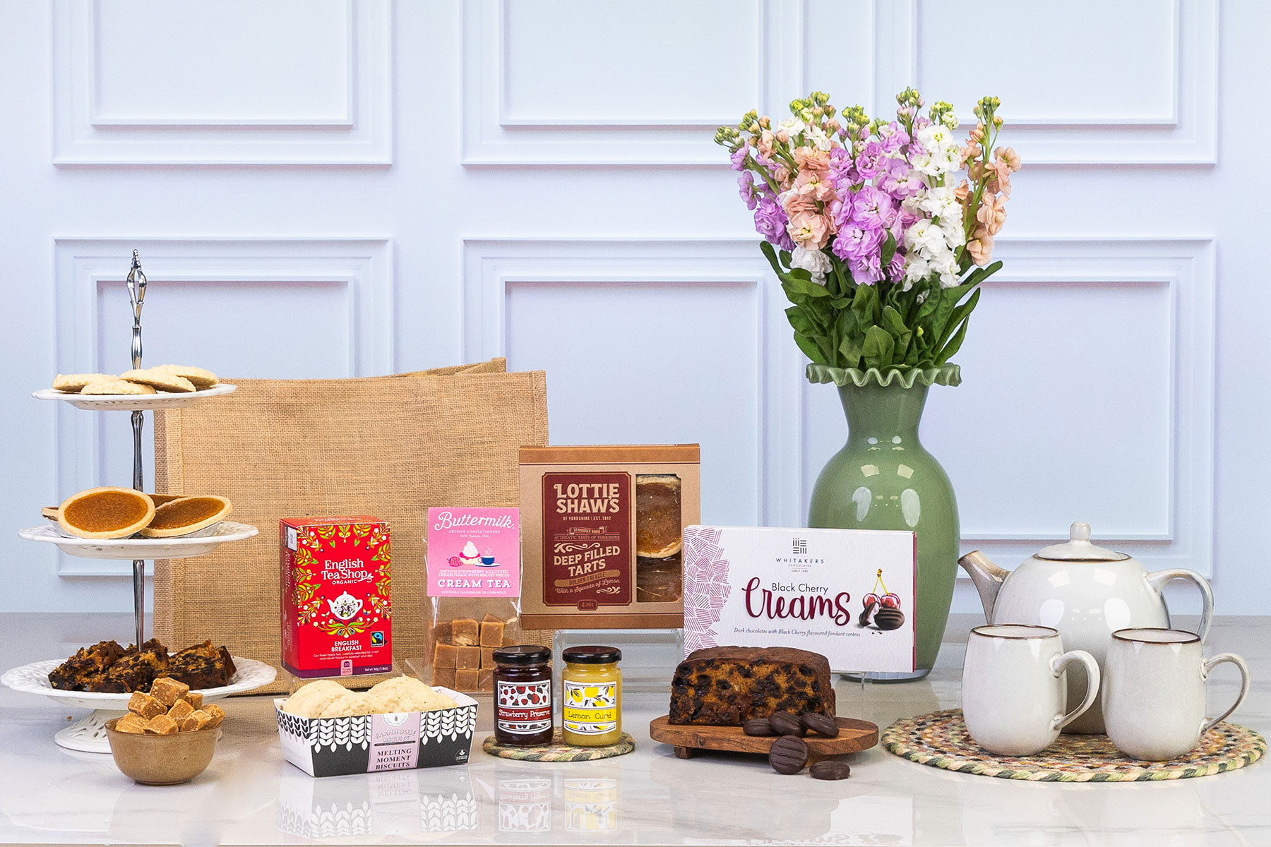 Traditional afternoon tea hamper featuring English Breakfast tea, fruit cake, deep-filled tarts, biscuits, cream tea fudge, preserves, cherry chocolates, teapot and mugs with fresh flowers.