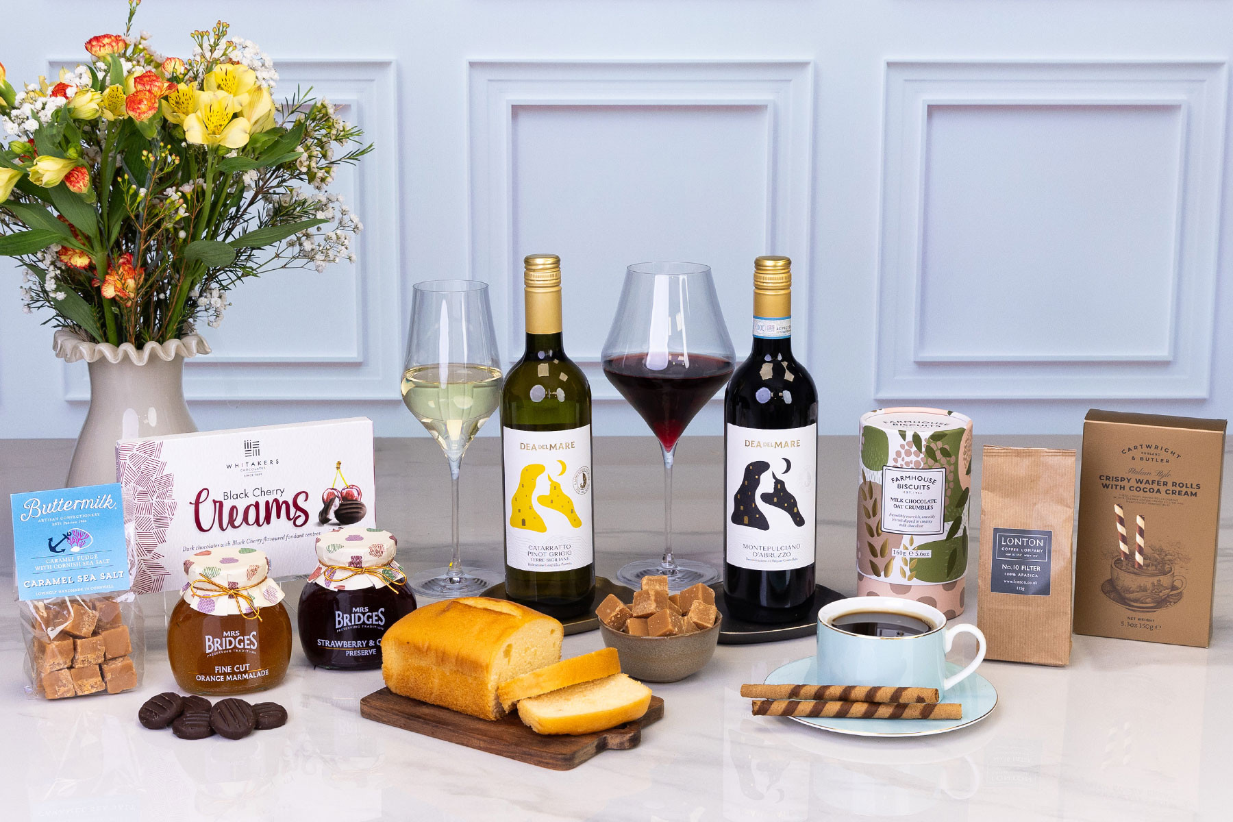 Luxury food and wine gift hamper featuring red and white wines, coffee, preserves, fudge, loaf cake, chocolates, biscuits and wafer rolls, displayed with fresh flowers.