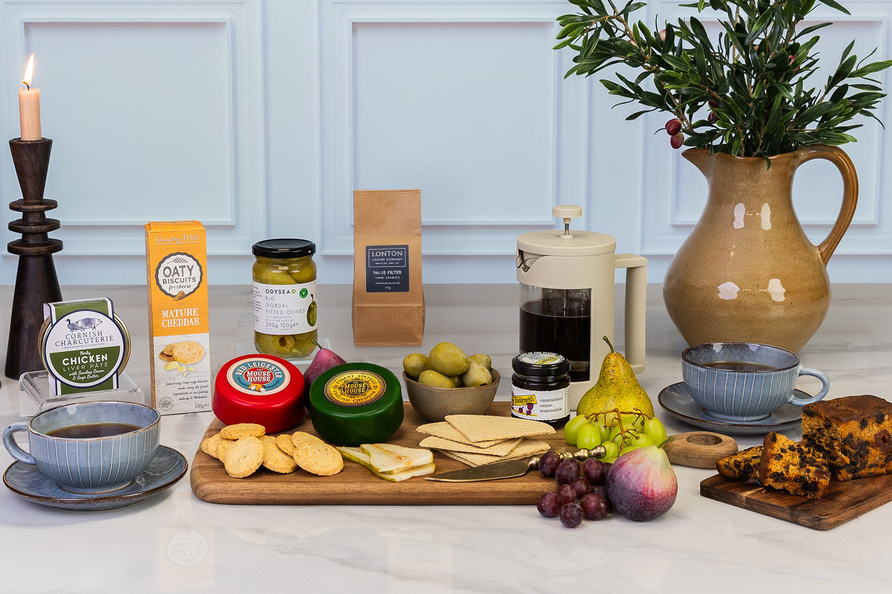 Luxury savoury food hamper featuring artisan cheeses, pâté, olives, coffee, oat biscuits, fruit cake, crackers, fruit and coffee served alongside a cafetière.