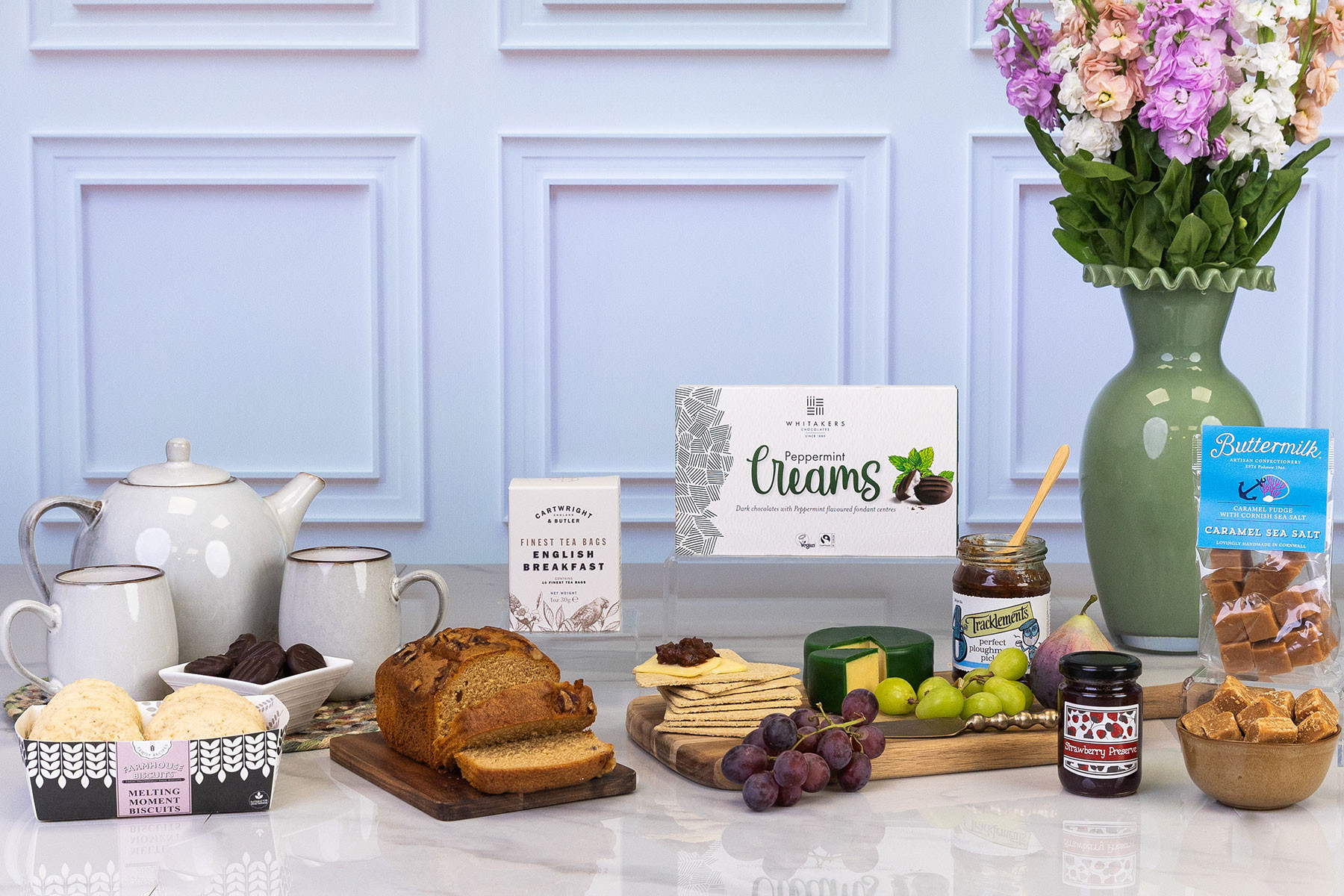 Luxury food and drink hamper featuring English breakfast tea, peppermint creams, caramel fudge, preserve, pickle, waxed cheese, artisan crackers, loaf cake, biscuits and teaware.