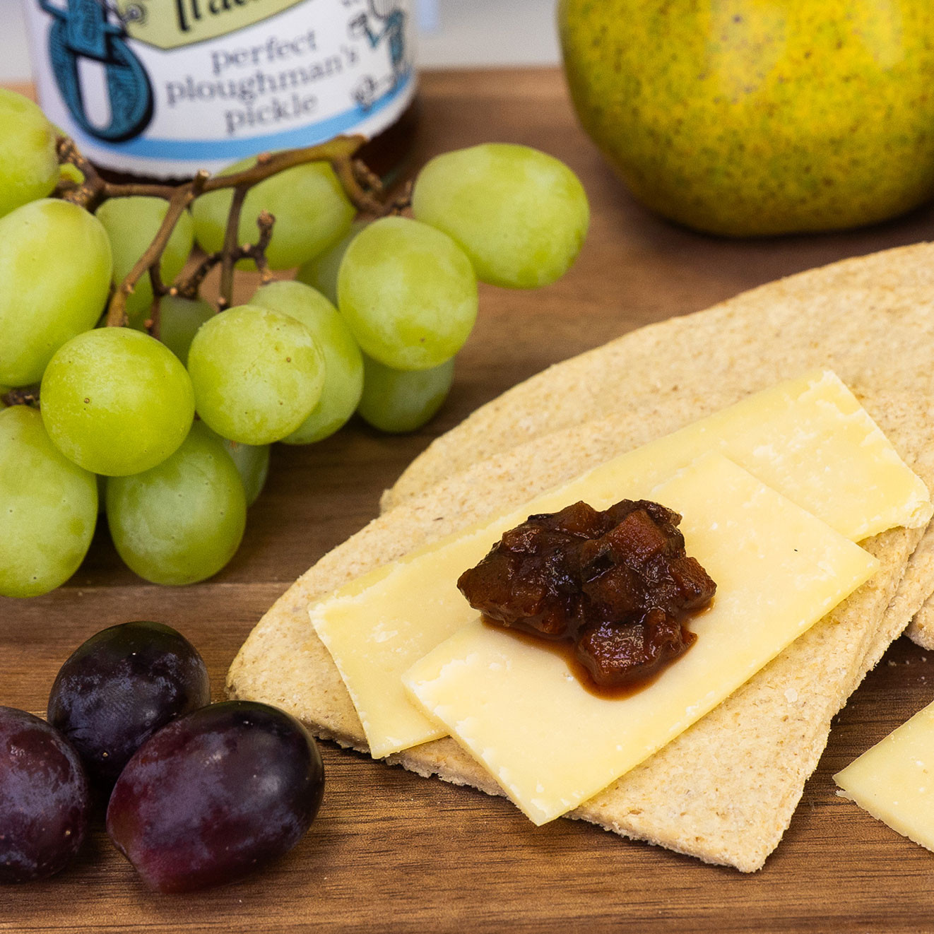 Artisan crackers topped with sliced cheese and Tracklements Ploughman's Pickle, served alongside fresh grapes and seasonal fruit.