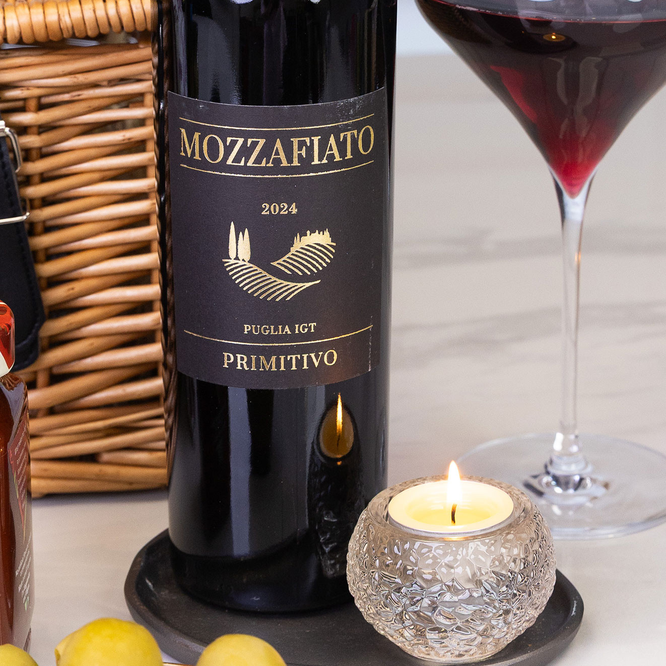 Bottle of Mozzafiato Primitivo Italian red wine beside a glass of red wine and candle, perfect for relaxed evenings and food pairings.