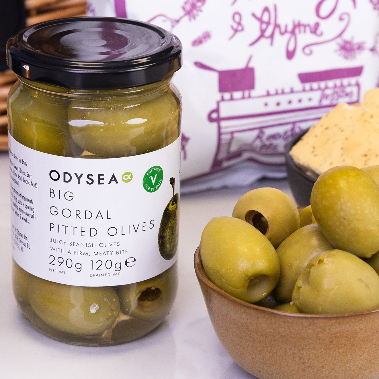 Jar of Odysea Big Gordal Pitted Olives served alongside a bowl of large Spanish green olives and artisan crackers.
