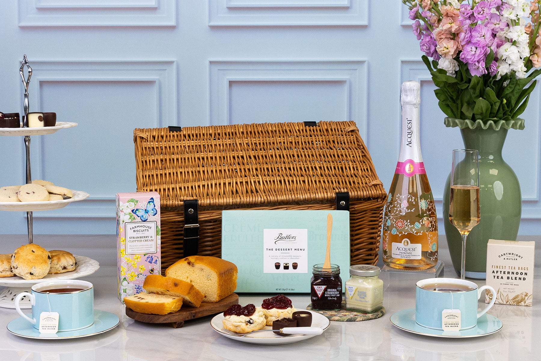 Luxury afternoon tea hamper with sparkling rosé, artisan tea, fruit loaf, chocolates, scones, preserves and clotted cream presented beside a traditional wicker picnic basket.