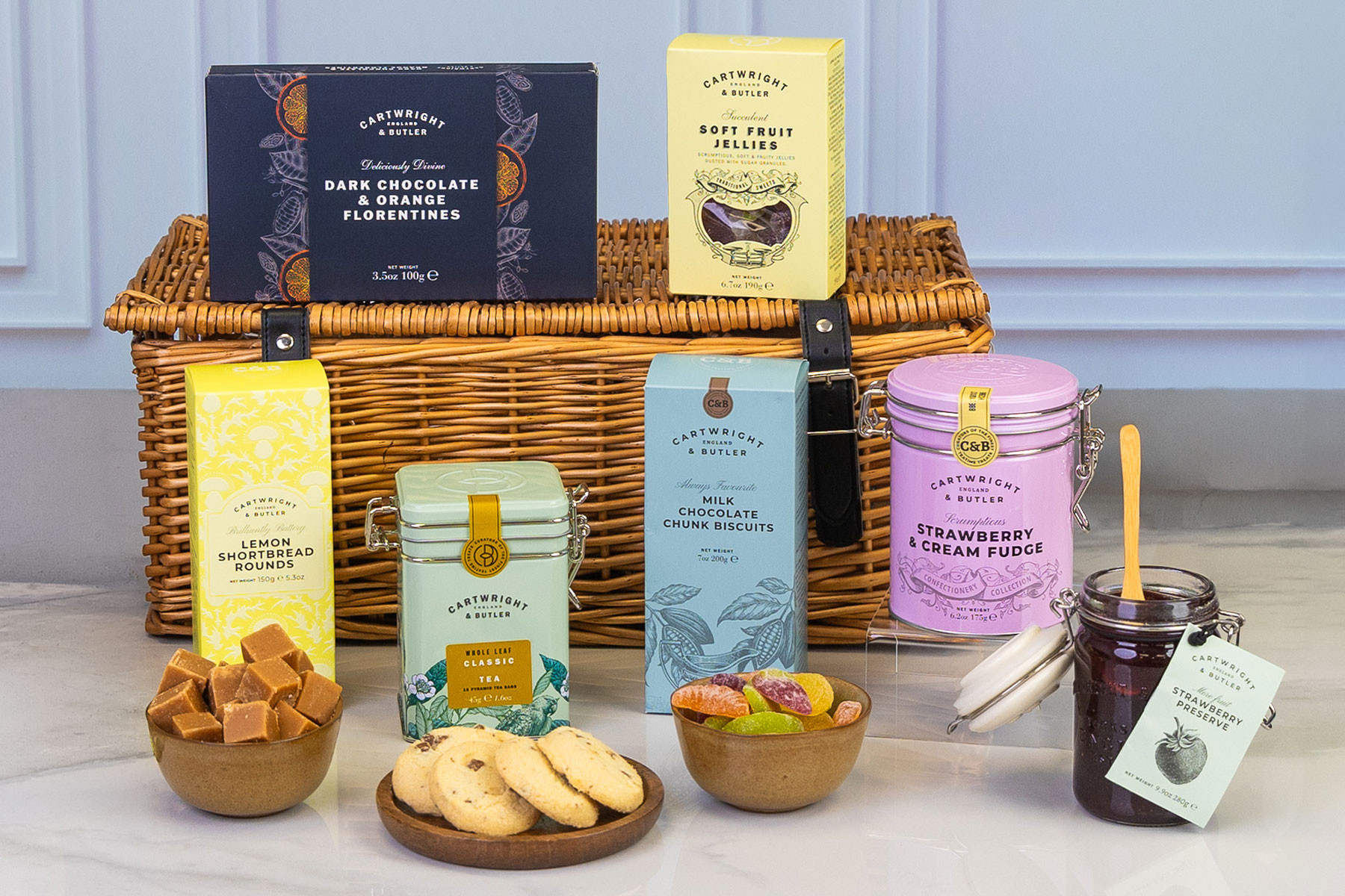 Luxury Cartwright & Butler gift hamper featuring artisan biscuits, tea, strawberry preserve, fruit jellies, fudge, Florentines and sweet treats presented in front of a traditional wicker picnic basket.