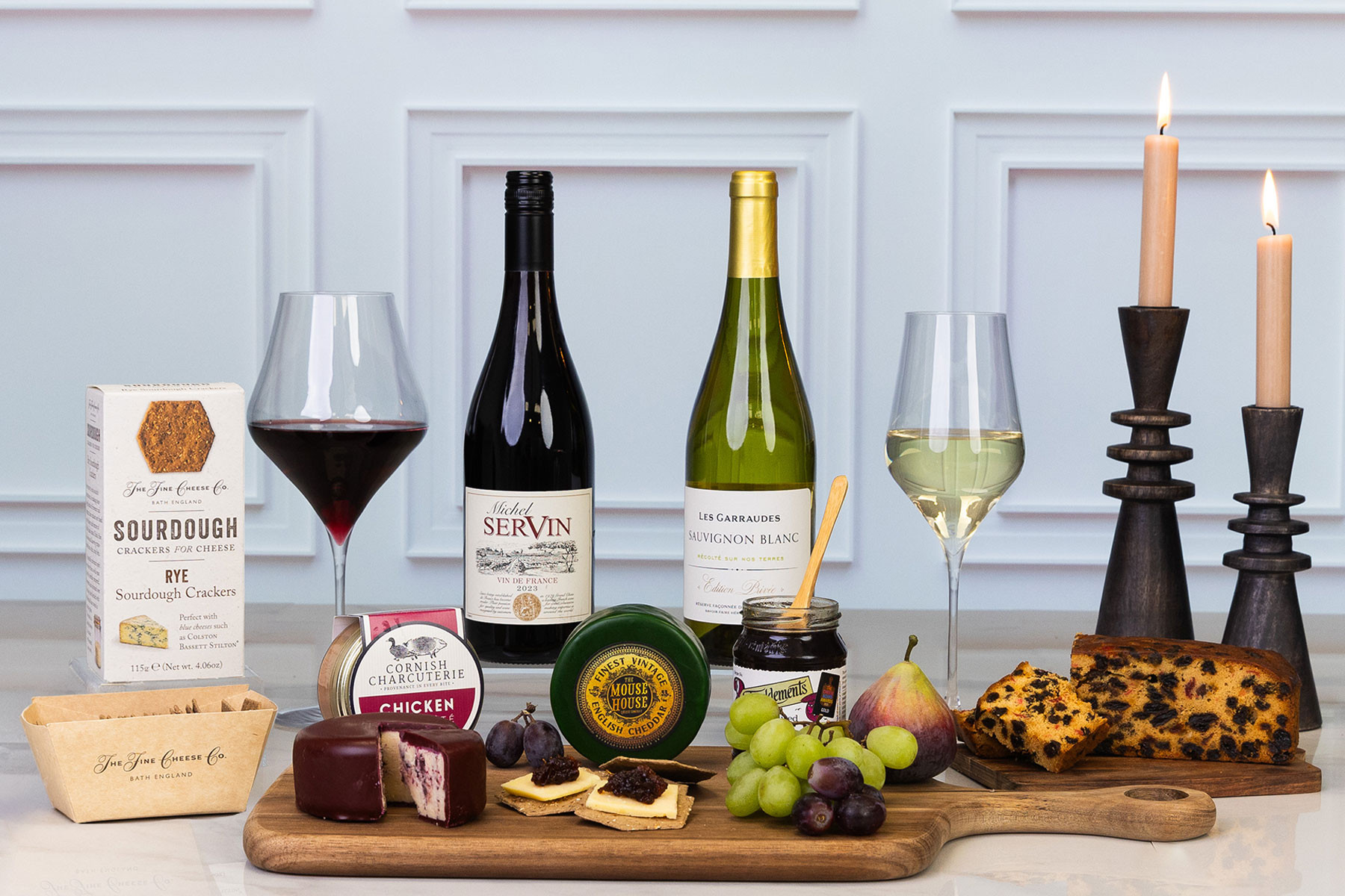 Luxury gourmet hamper featuring artisan cheeses, chicken liver pâté, sourdough crackers, caramelised onion marmalade, rich fruit cake, premium red and white wine, and fresh fruit, elegantly displayed with lit candles.