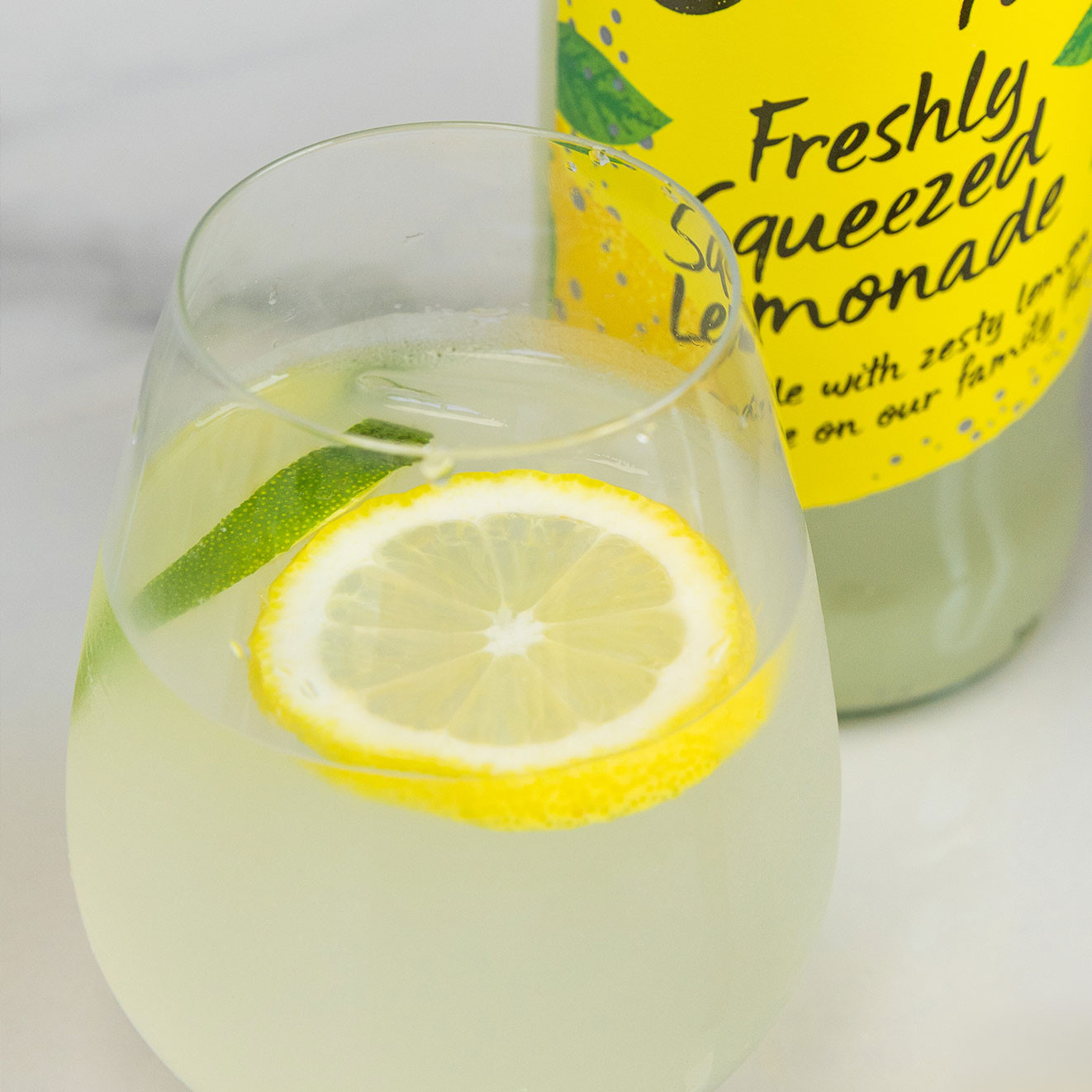 Glass of freshly squeezed lemonade garnished with lemon and lime slices, served beside a bottle of Belvoir Farm Freshly Squeezed Lemonade.
