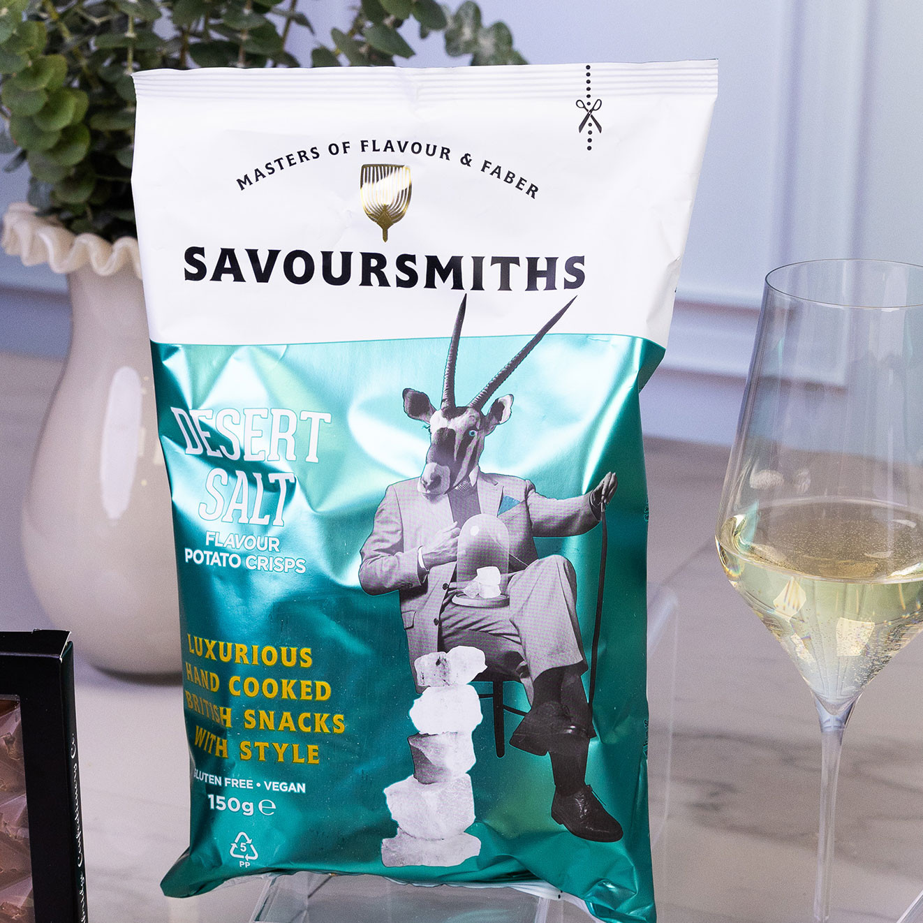 Bag of Savoursmiths Desert Salt hand-cooked potato crisps displayed beside a glass of white wine, highlighting a premium British savoury snack.