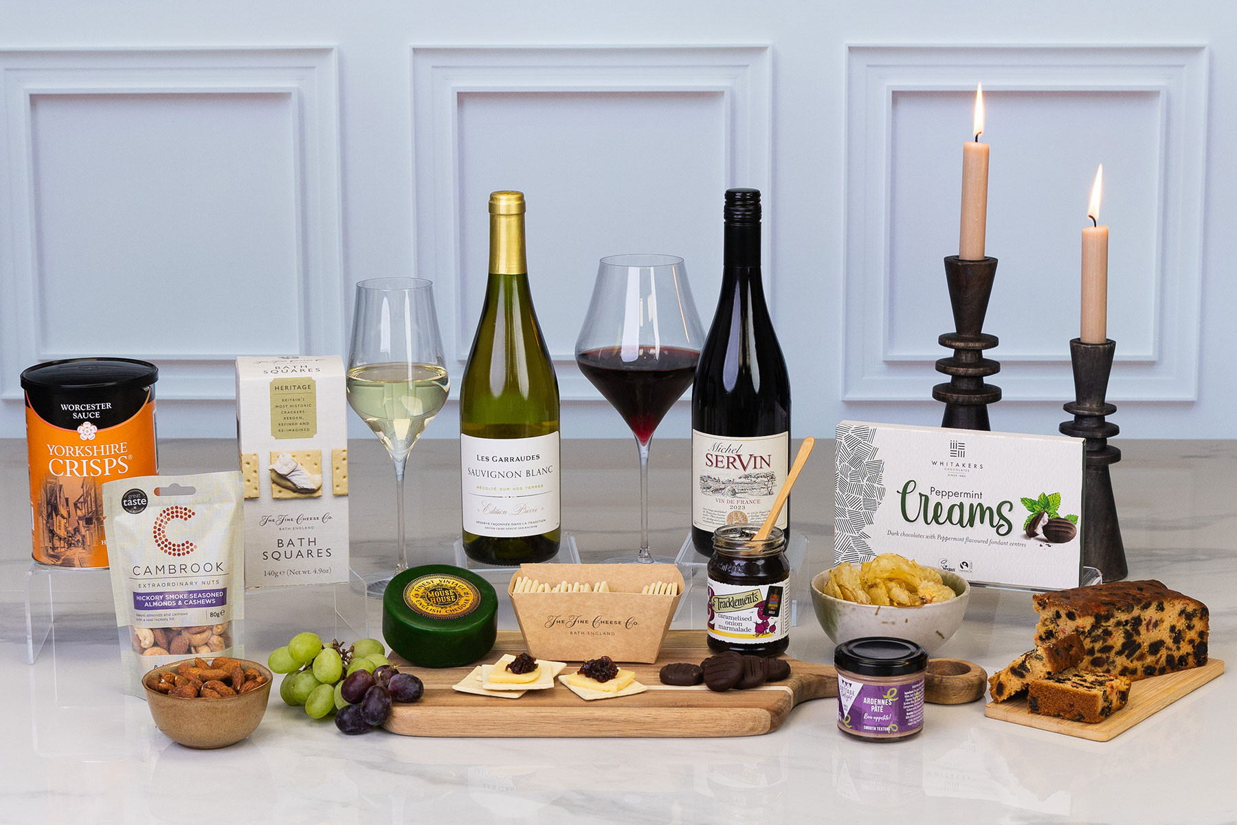 Luxury food and wine hamper featuring French Sauvignon Blanc, red wine, artisan cheeses, pâté, chutney, crackers, crisps, fruit cake, chocolates and gourmet snacks, elegantly displayed with candlelight.