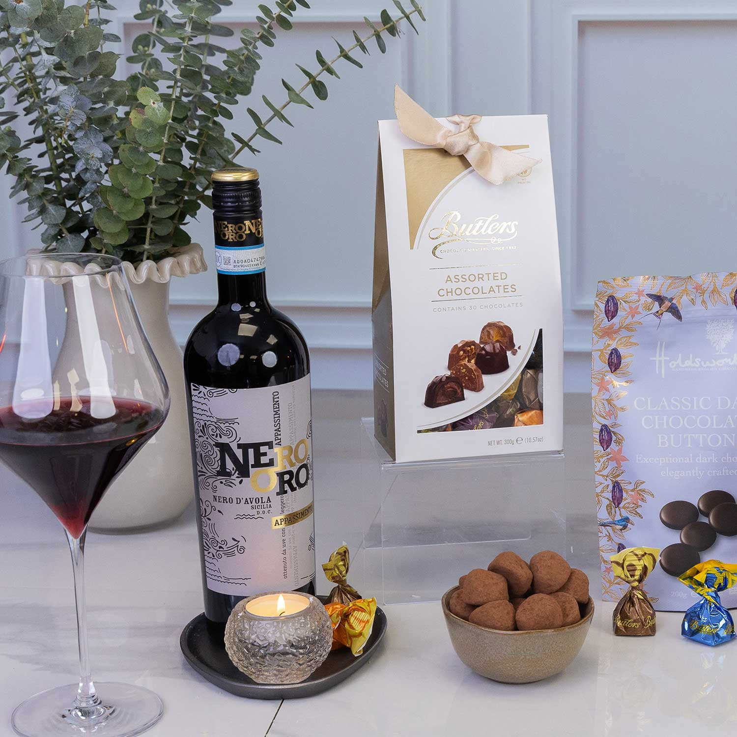 Close-up of the Luxury wine and chocolate gift hamper featuring Nero d'Avola red wine, premium chocolates, chocolate-coated Brazil nuts, hazelnut truffles, artisan chocolate bars and assorted chocolate selection, elegantly presented with candlelight.