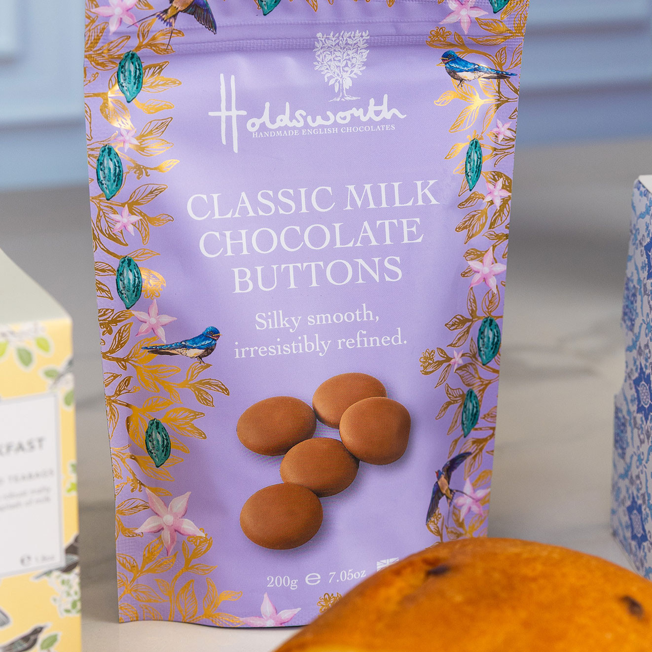 Holdsworth Classic Milk Chocolate Buttons in decorative lilac packaging with floral and bird illustrations, displayed as part of an elegant tea and chocolate gift selection.