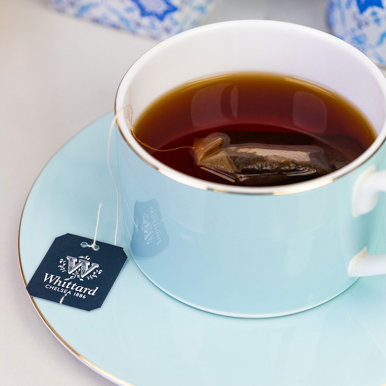 Close-up of a pale blue teacup and saucer filled with Earl Grey tea, with a Whittard of Chelsea teabag steeping in the cup and branded tag resting on the saucer.