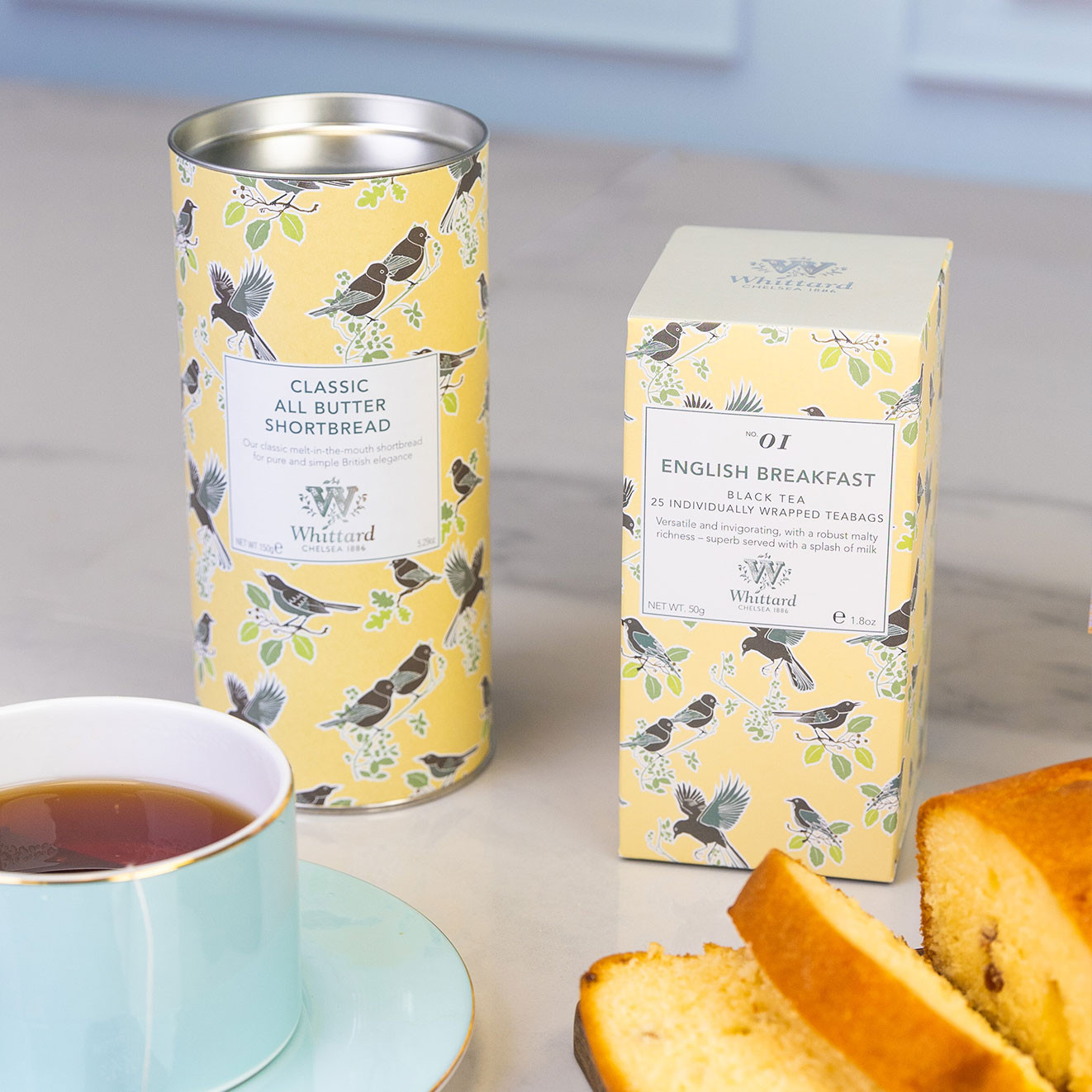 Whittard of Chelsea English Breakfast black tea and Classic All Butter Shortbread in coordinating yellow bird-patterned packaging, styled with a pale blue cup of tea and slices of fruit cake.