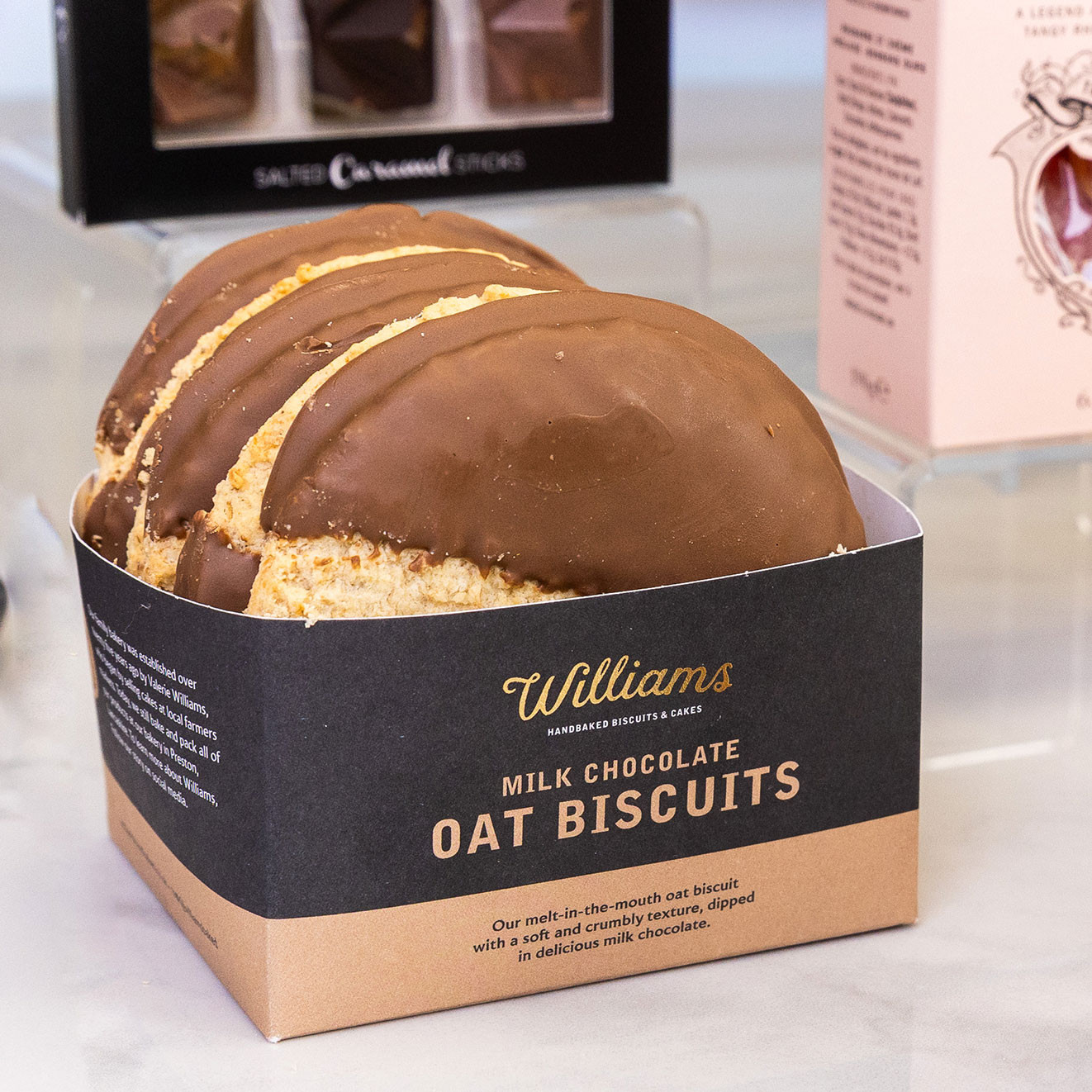 Close-up of Williams milk chocolate oat biscuits displayed in an open box, with chocolate and confectionery products in the background.