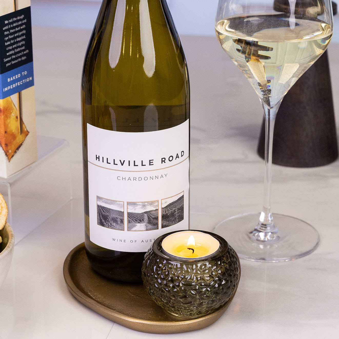 Close-up of a bottle of Hillville Road Chardonnay beside a glass of white wine and a lit candle on a tabletop.