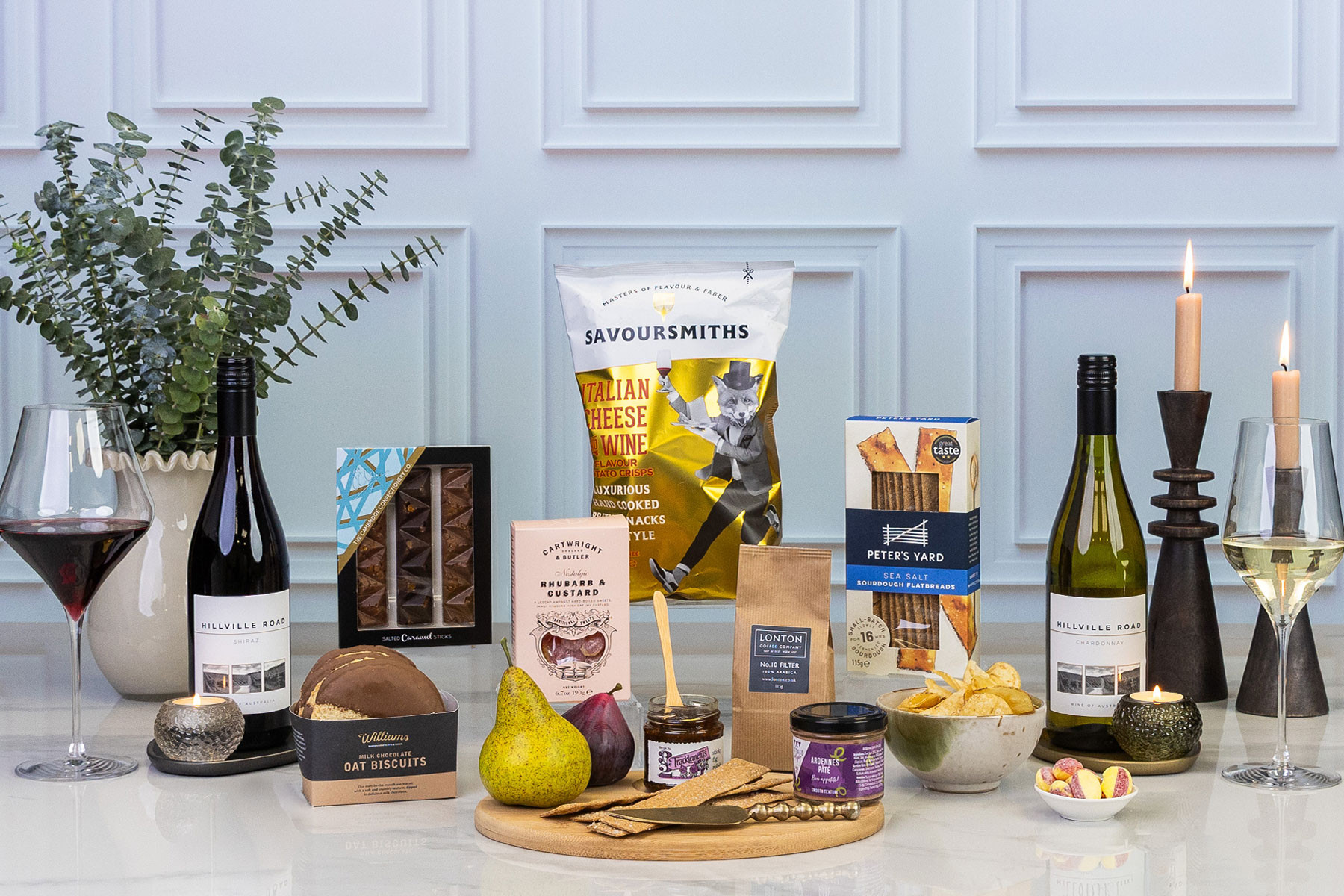 Gourmet gift selection featuring Hillville Road red and white wines, chocolates, biscuits, coffee, crackers, crisps, preserves and fresh fruit, styled with wine glasses and lit candles.