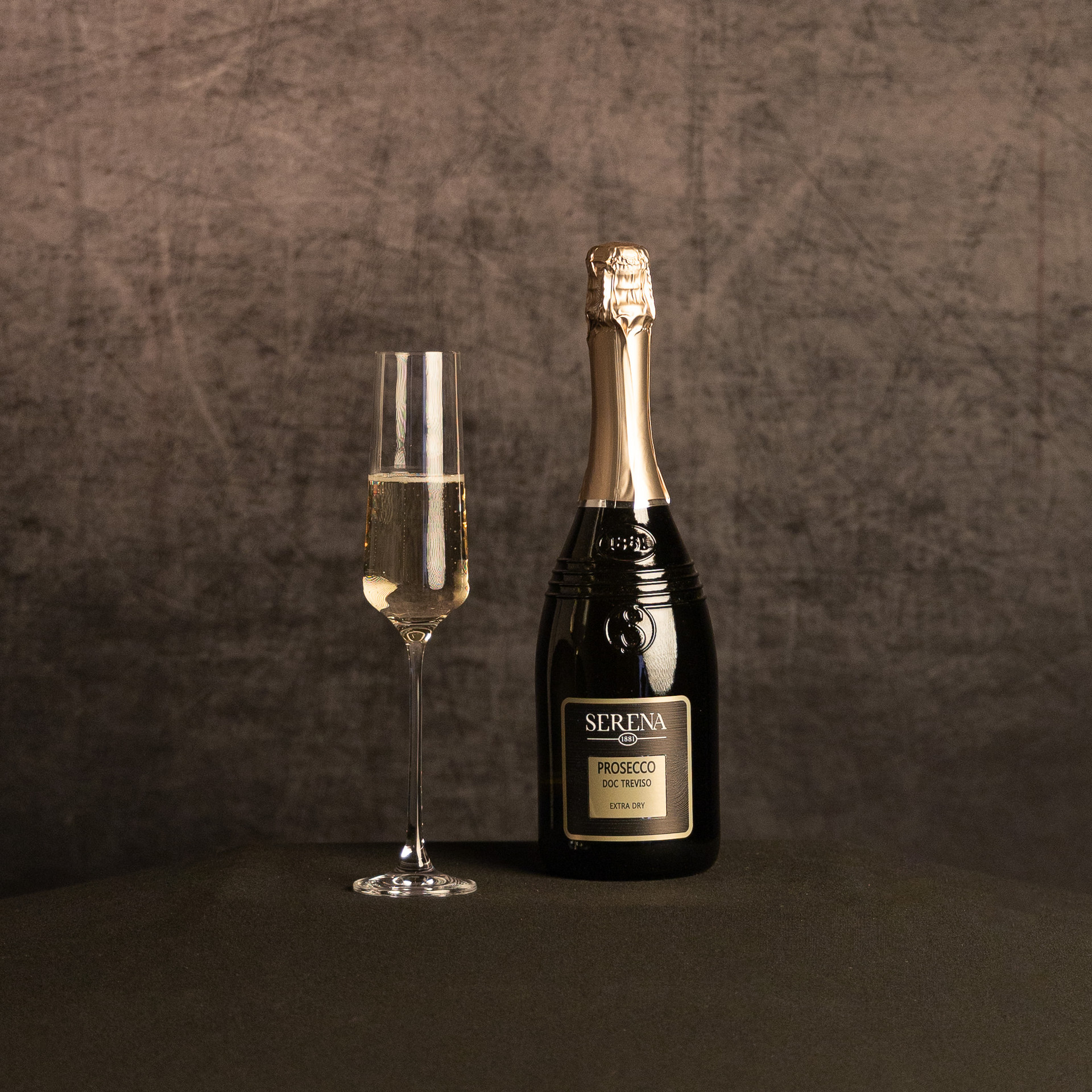 A bottle of Serena 1881 Prosecco DOC Treviso Extra Dry beside a partially filled champagne flute, photographed against a dark, textured brown backdrop.