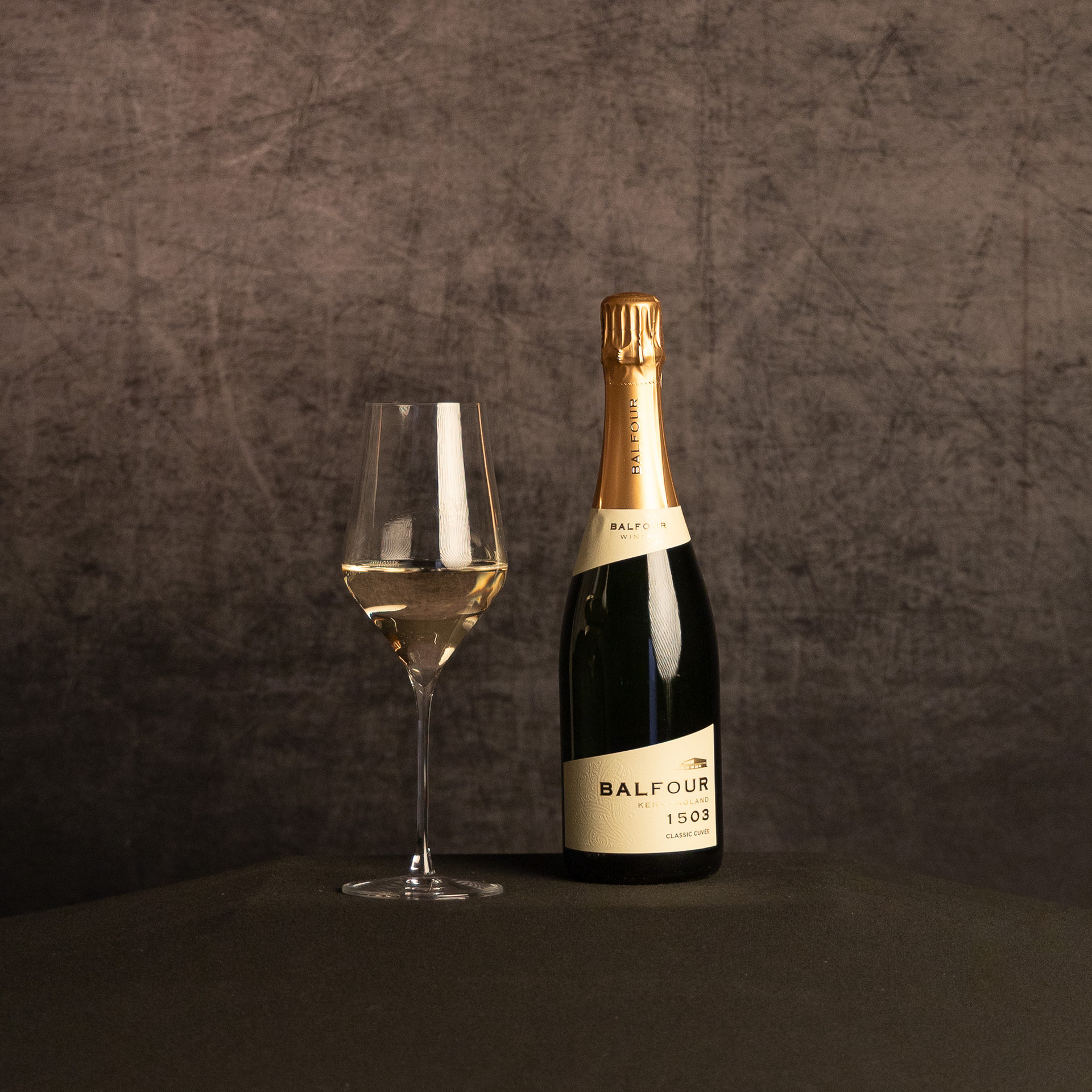 A bottle of Balfour 1503 Classic Cuvée sparkling wine beside a partially filled wine glass, photographed against a dark, textured brown backdrop.