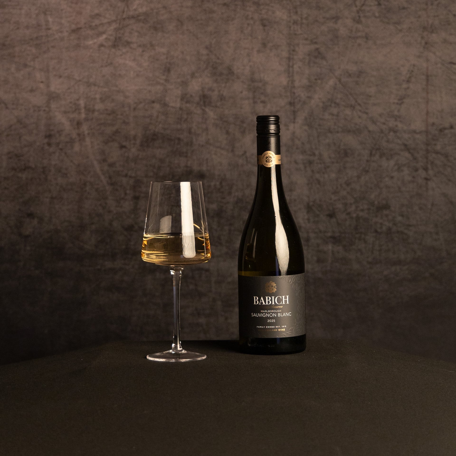 A bottle of Babich Reserve Marlborough Sauvignon Blanc beside a partially filled wine glass, photographed against a dark, textured brown backdrop.