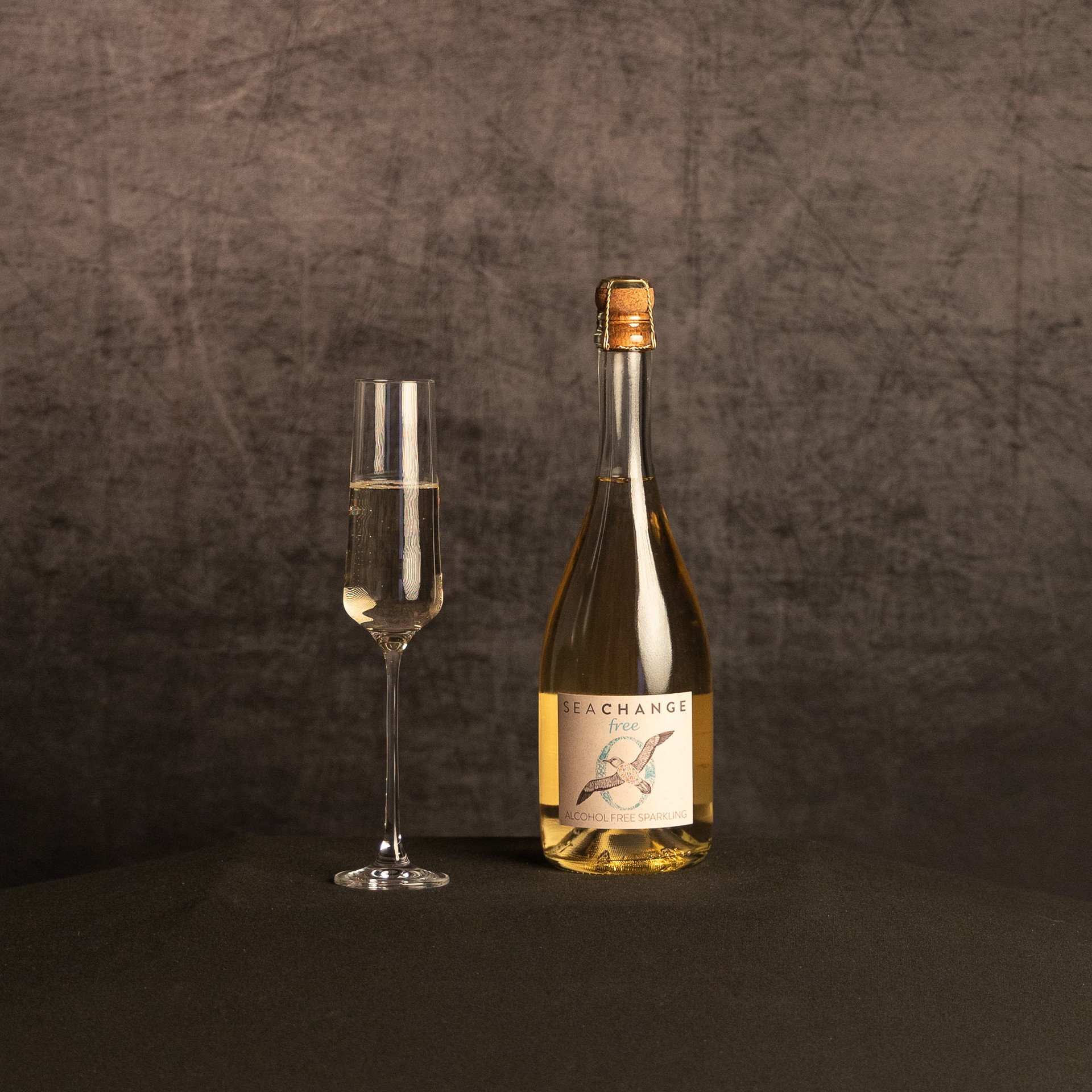 A bottle of Sea Change Free alcohol-free sparkling wine beside a partially filled champagne flute, photographed against a dark, textured brown backdrop.