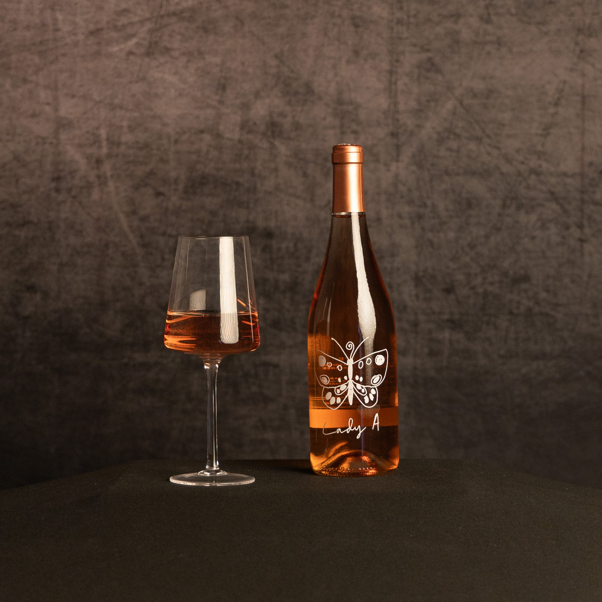 A bottle of Lady A rosé beside a partially filled glass of rosé wine, photographed against a dark, textured brown backdrop.