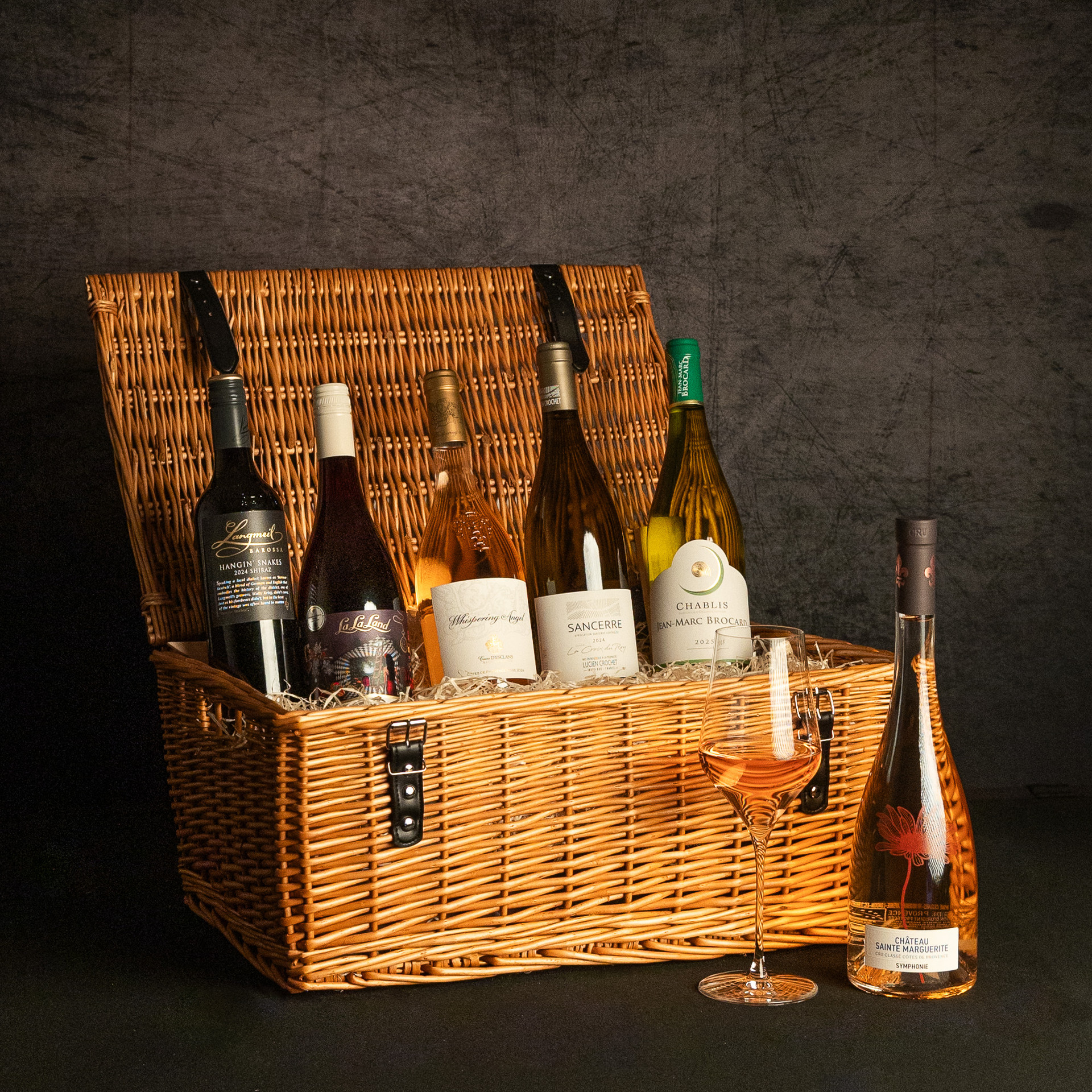 An open wicker hamper filled with five assorted wines, including Shiraz, Pinot Noir, rosé, Sancerre and Chablis, with a glass of rosé and a Château Sainte Marguerite Symphonie rosé bottle displayed alongside.