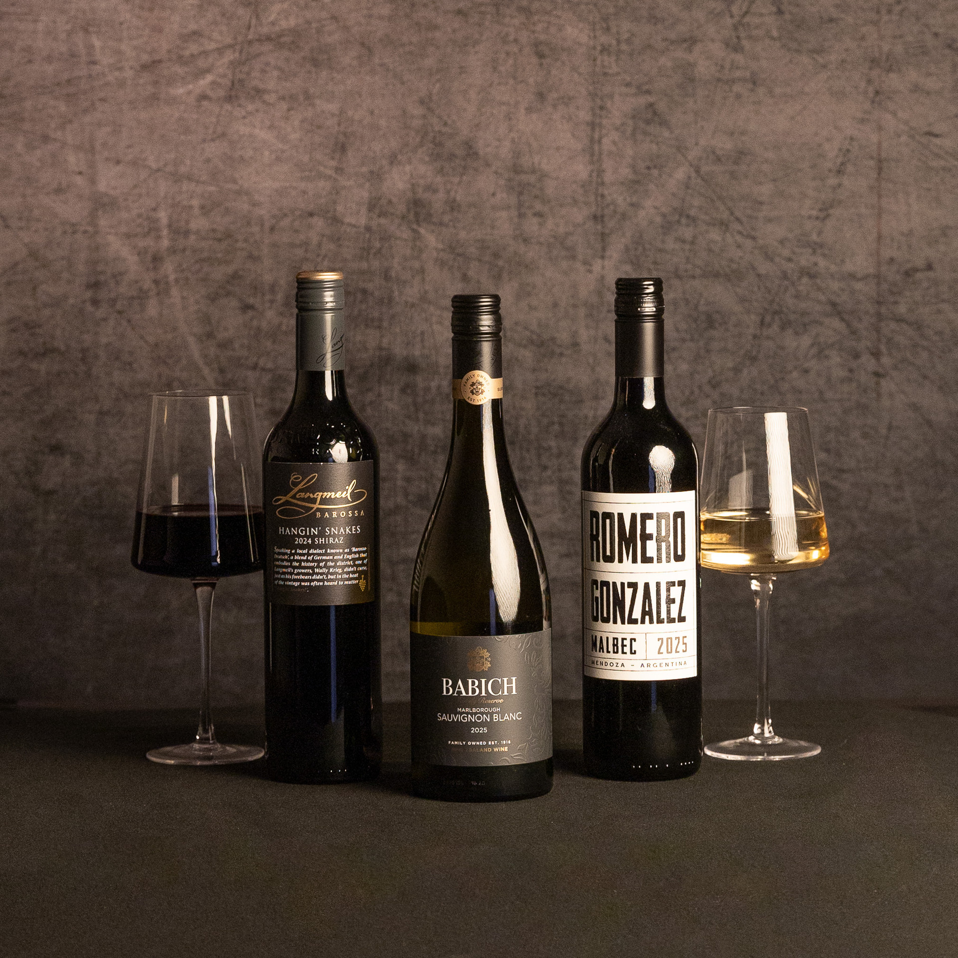 Three wine bottles—Langmeil Hangin’ Snakes Shiraz, Babich Marlborough Sauvignon Blanc and Romero Gonzalez Malbec—are displayed between glasses of red and white wine against a dark, textured backdrop.