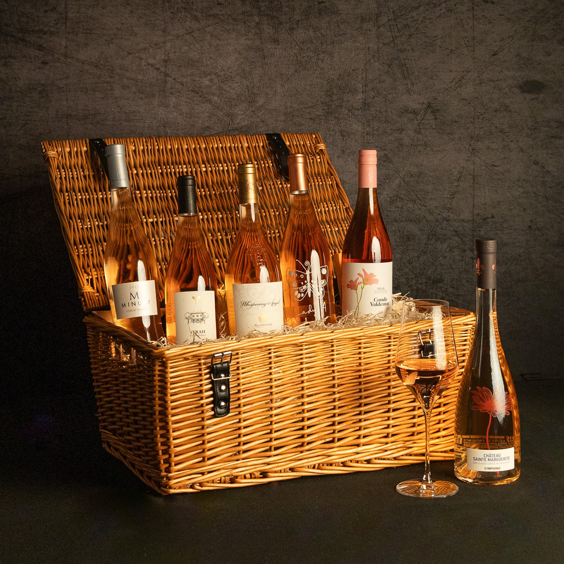 An open wicker hamper filled with five bottles of rosé wine, with a glass of rosé and a Château Sainte Marguerite Symphonie rosé bottle displayed alongside.