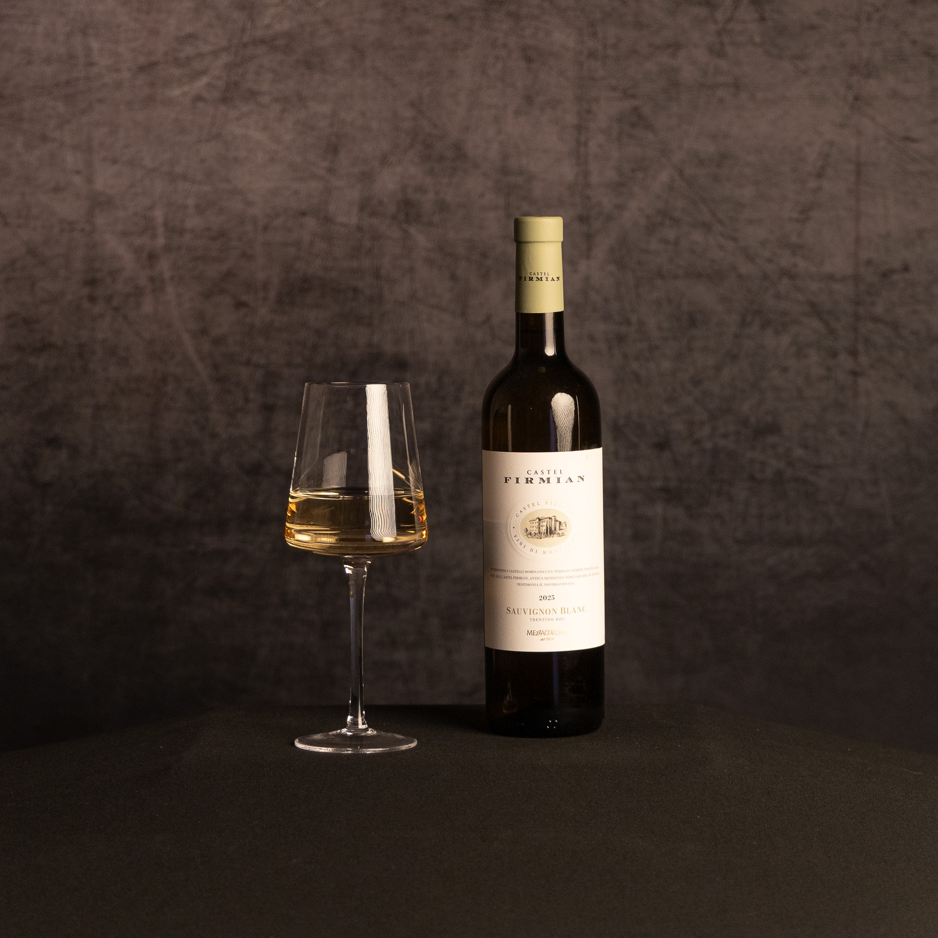 A bottle of Castel Firmian Sauvignon Blanc beside a partially filled wine glass, photographed against a dark, textured brown backdrop.