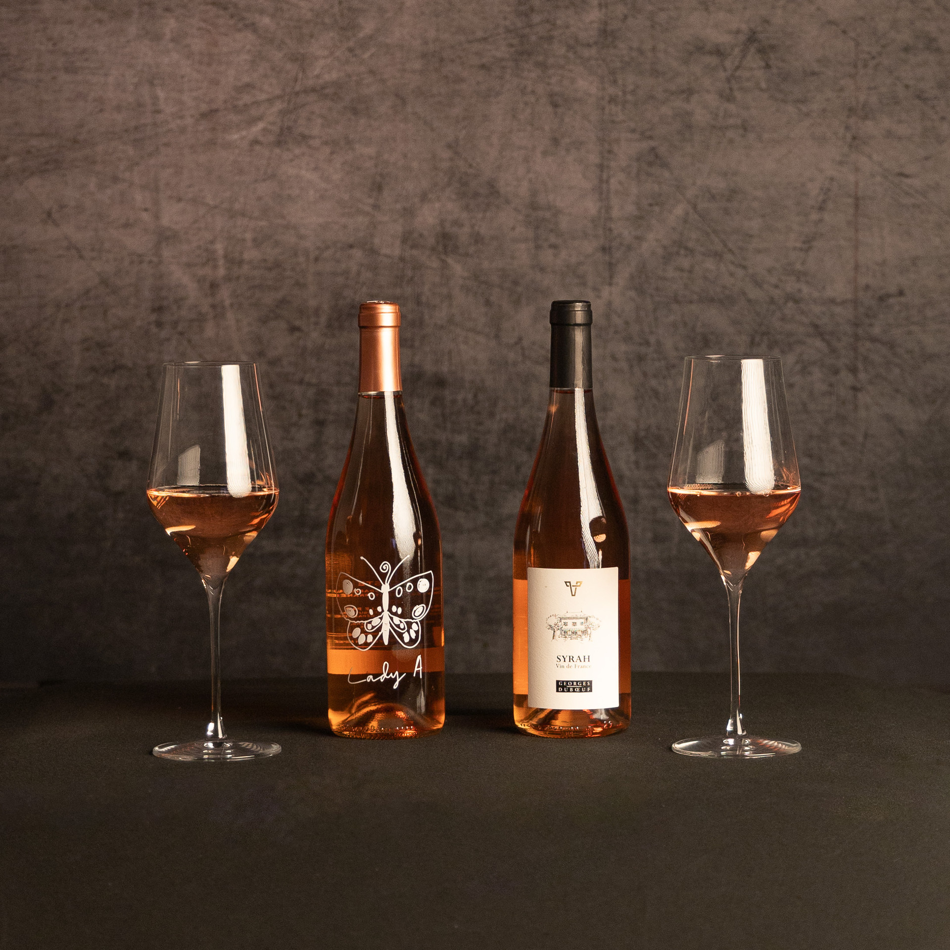 Bottles of Lady A rosé and Georges Duboeuf Syrah rosé stand between two glasses of pink wine against a dark, textured backdrop.