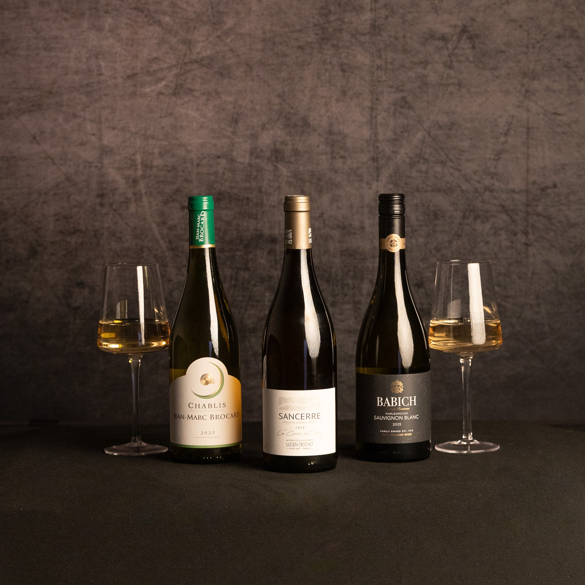 Three white wine bottles—Jean-Marc Brocard Chablis, Lucien Crochet Sancerre and Babich Marlborough Sauvignon Blanc—are displayed between two glasses of white wine against a dark, textured backdrop.