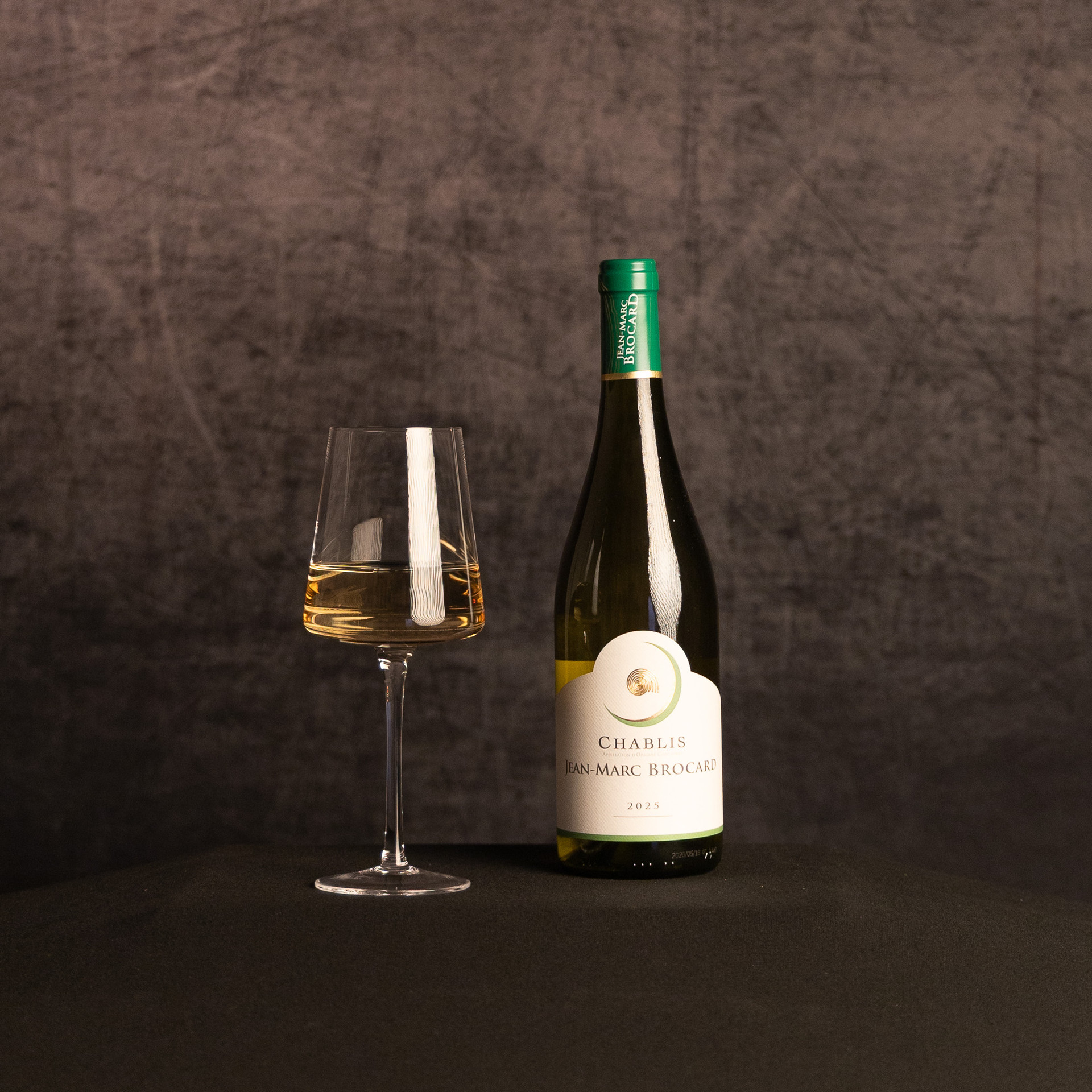 A bottle of Jean-Marc Brocard Chablis beside a partially filled wine glass, photographed against a dark, textured brown backdrop.