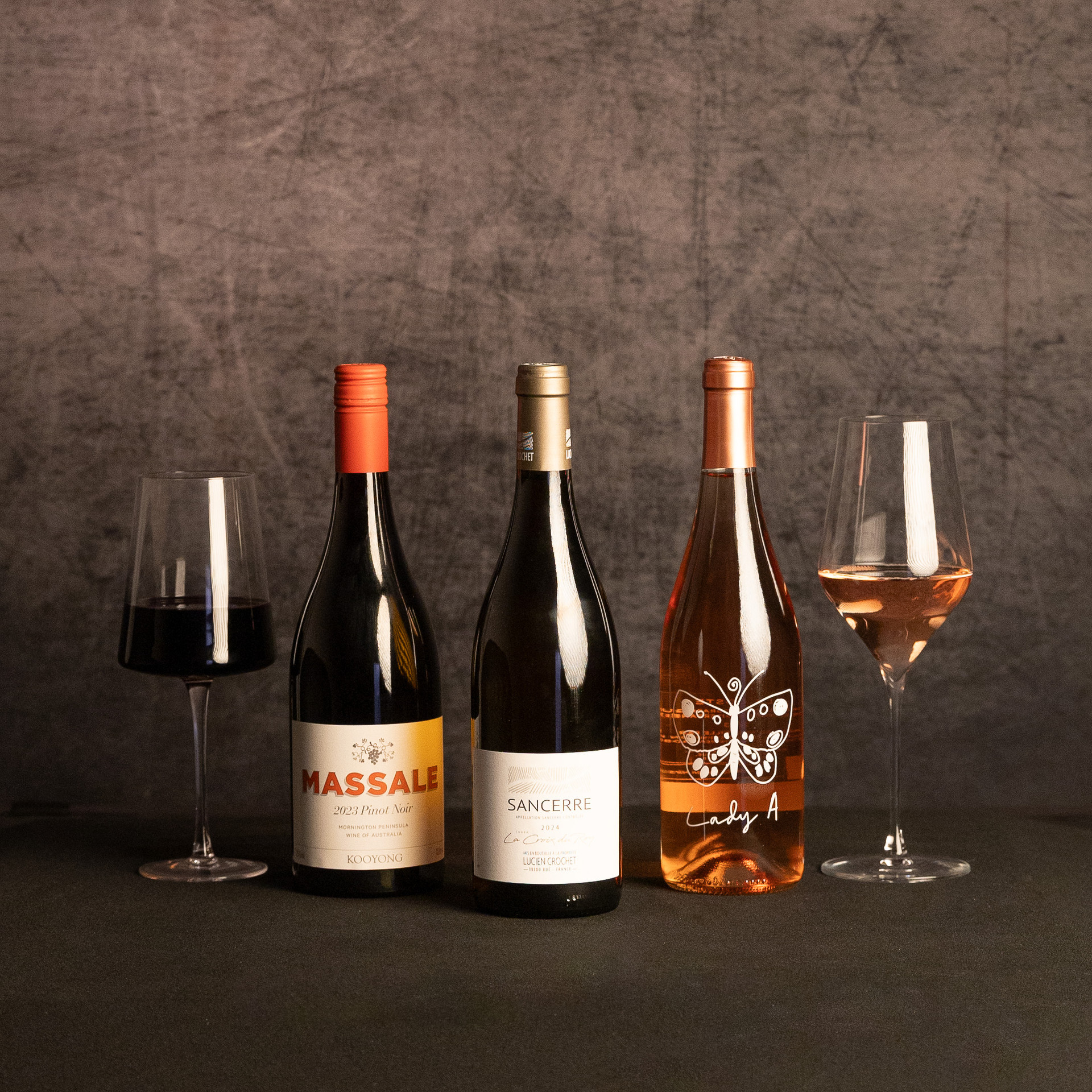 Three wine bottles—Massale Pinot Noir, Lucien Crochet Sancerre and Lady A rosé—are displayed between glasses of red and rosé wine against a dark, textured backdrop.