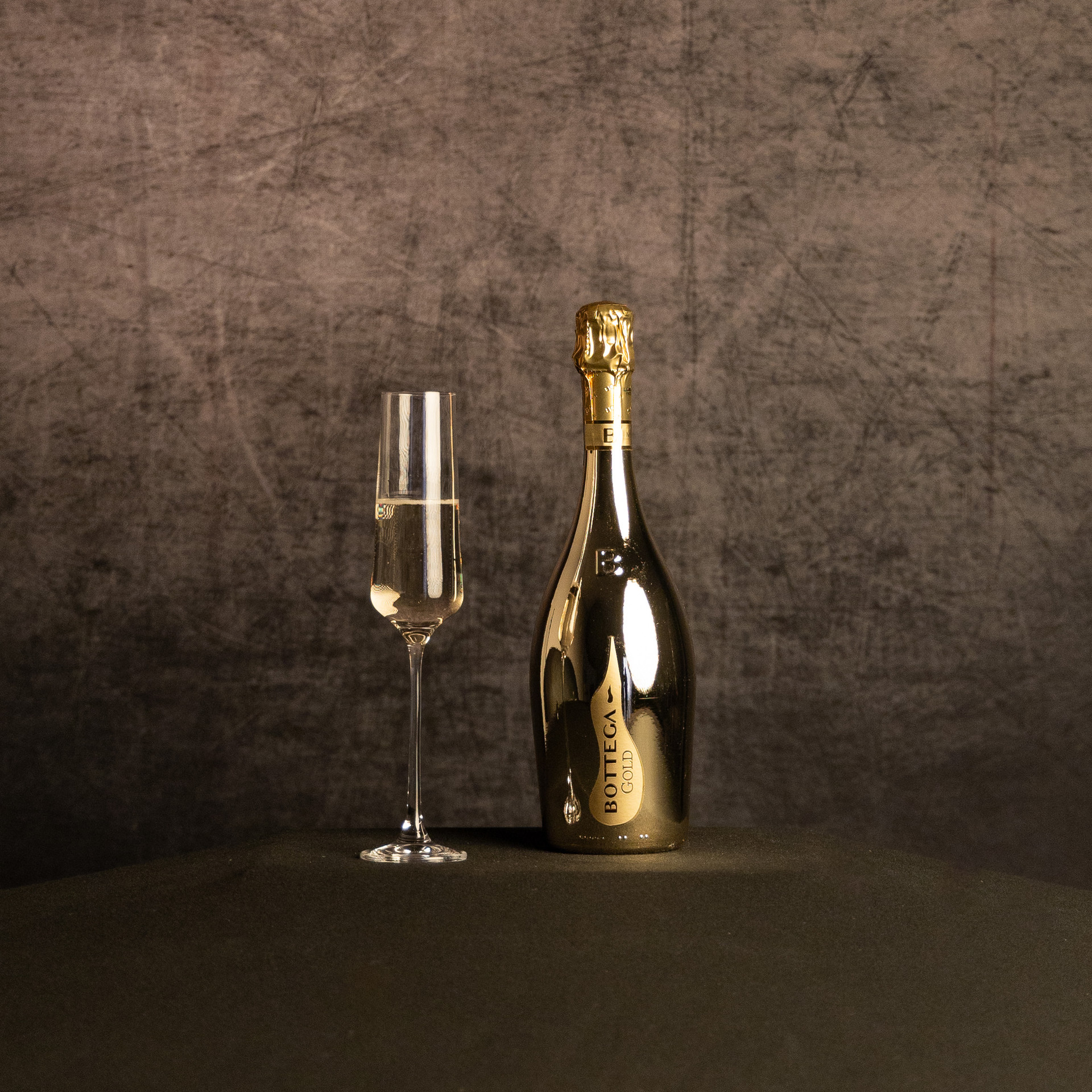 A bottle of Bottega Gold Prosecco beside a partially filled champagne flute, photographed against a dark, textured brown backdrop.