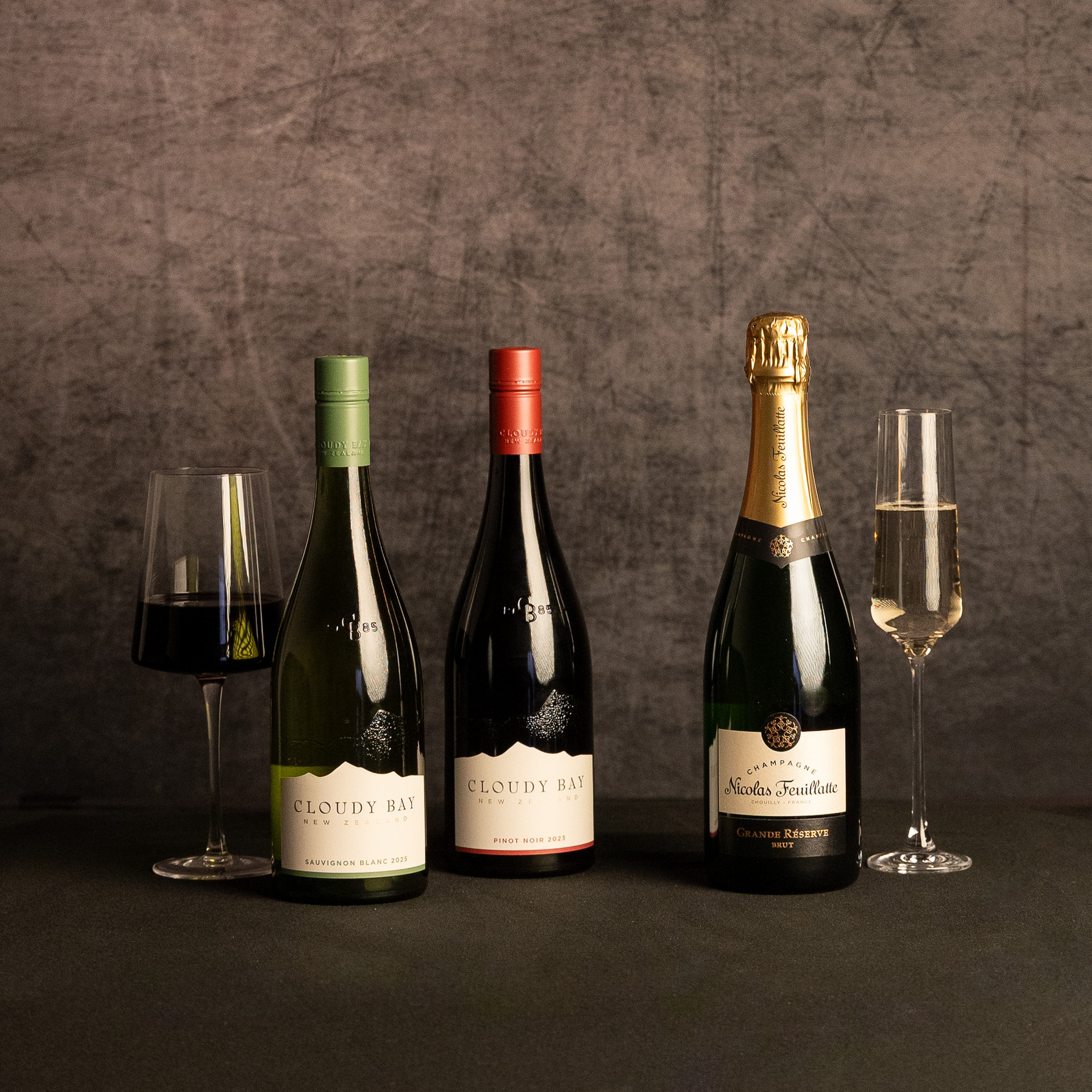 Three bottles—Cloudy Bay Sauvignon Blanc, Cloudy Bay Pinot Noir and Nicolas Feuillatte Grande Réserve Brut Champagne—are displayed between glasses of red and sparkling wine against a dark, textured backdrop.