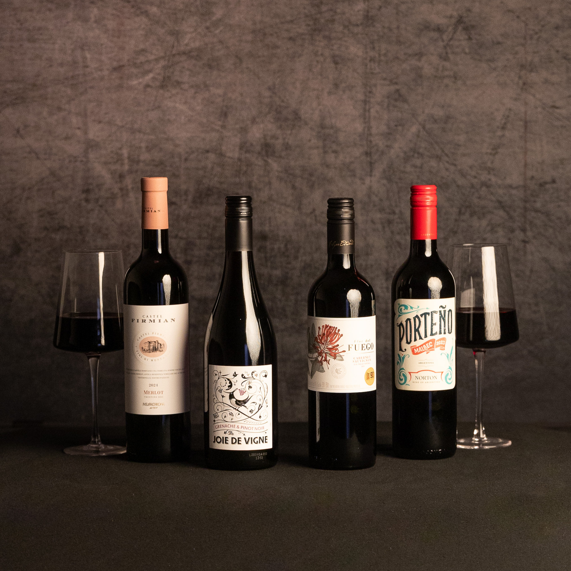 Four red wine bottles—Castel Firmian Merlot, Joie de Vigne Grenache & Pinot Noir, Flor del Fuego Cabernet Sauvignon and Porteño Malbec—are displayed between two glasses of red wine against a dark, textured backdrop.