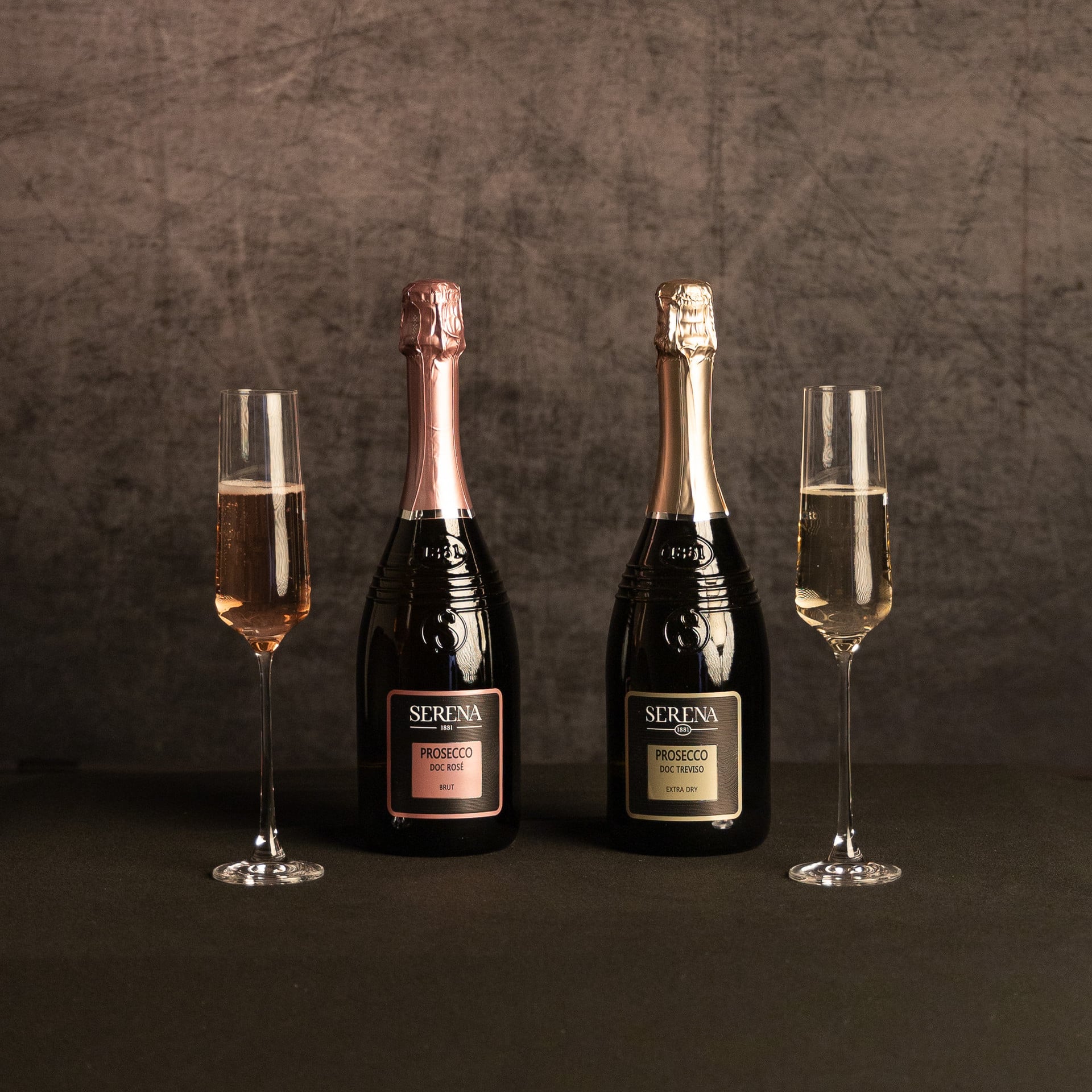 Two bottles of Serena Prosecco—DOC Rosé Brut and DOC Treviso Extra Dry—displayed between matching flutes of pink and pale sparkling wine against a dark, textured backdrop.