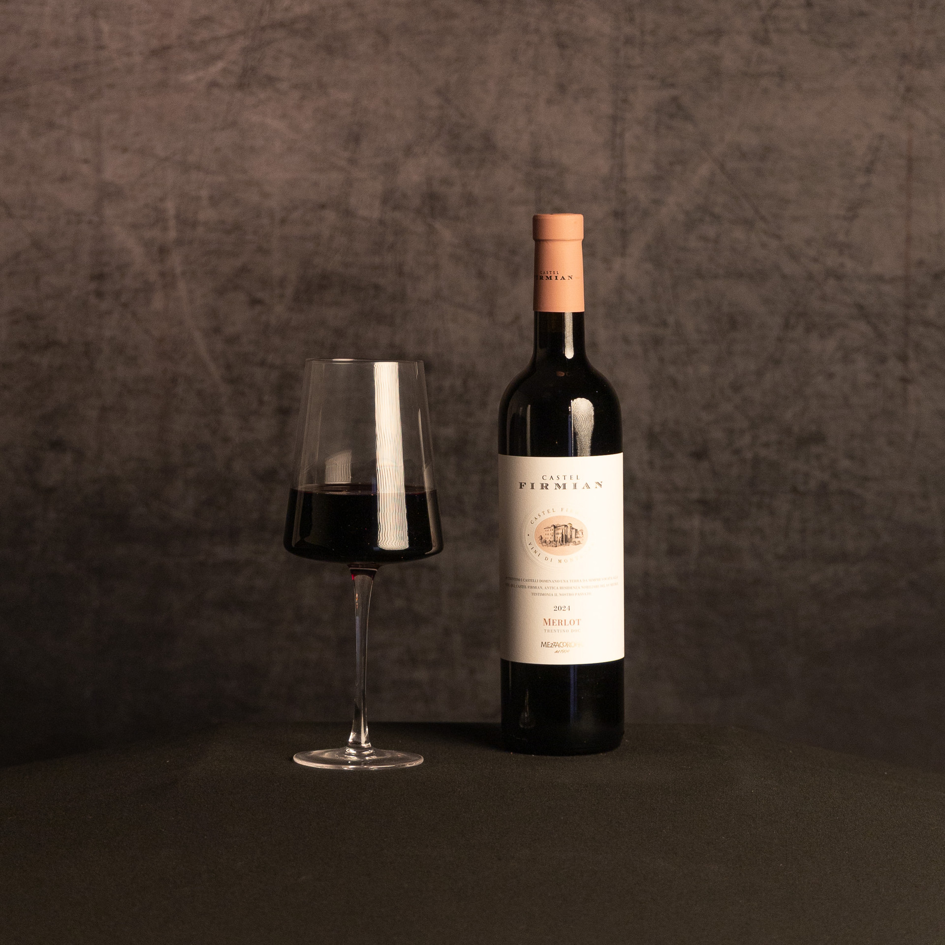A bottle of Castel Firmian Merlot beside a partially filled glass of red wine, photographed against a dark, textured brown backdrop.