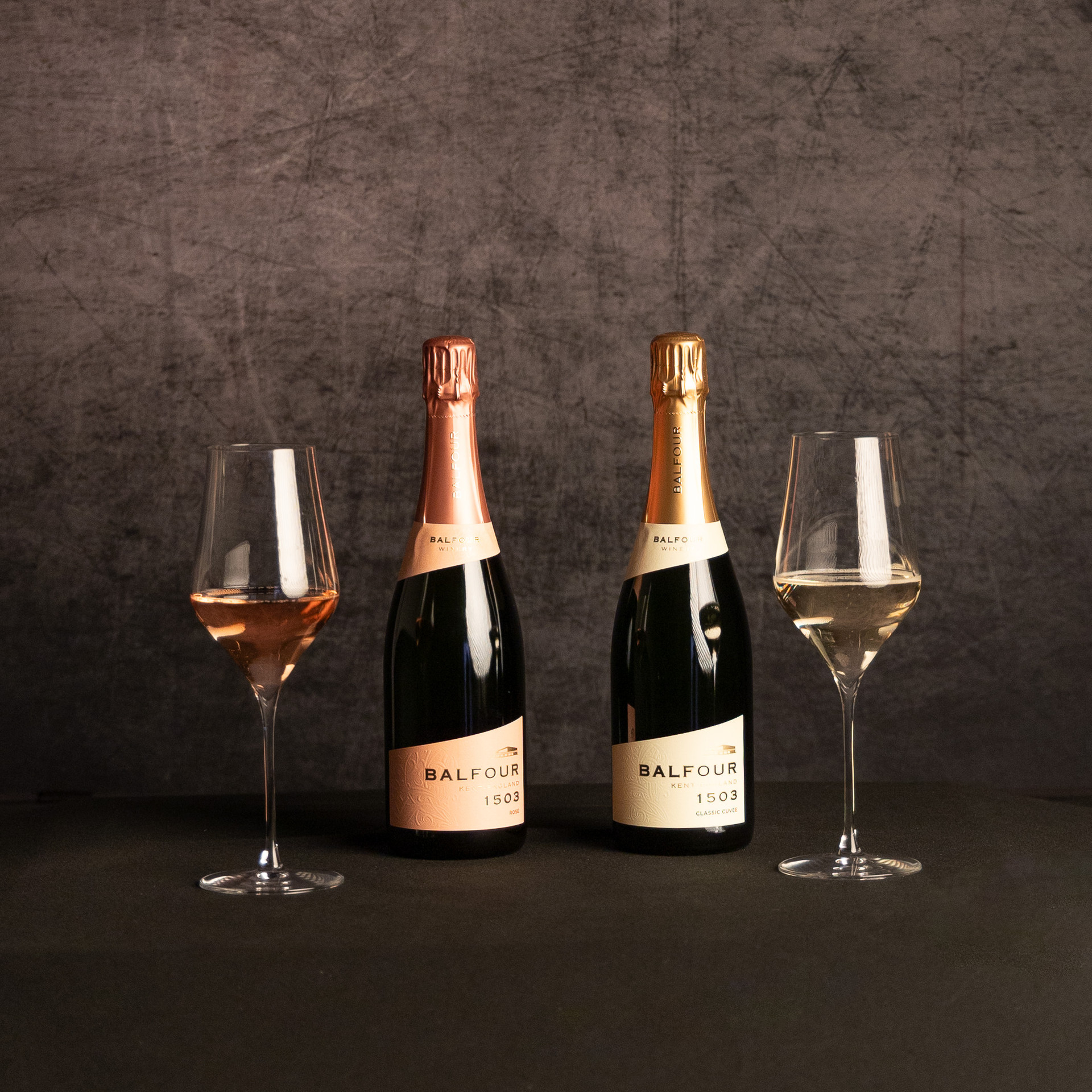 Two bottles of Balfour 1503 English sparkling wine—Rosé and Classic Cuvée—displayed between matching glasses of pink and pale sparkling wine against a dark, textured backdrop.