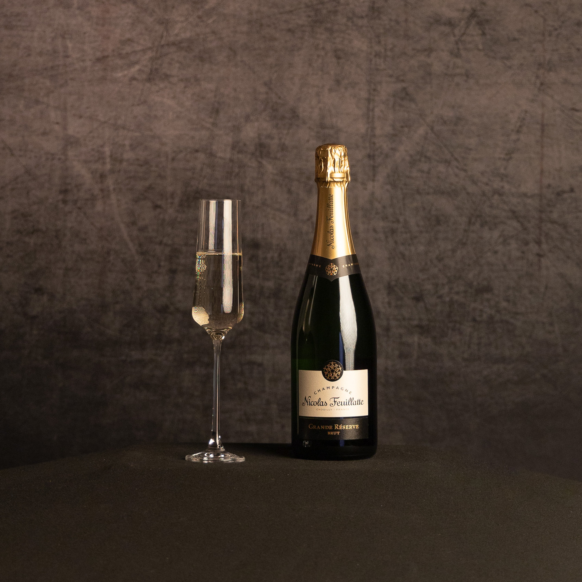 A bottle of Nicolas Feuillatte Grande Réserve Brut Champagne beside a partially filled champagne flute, photographed against a dark, textured brown backdrop.