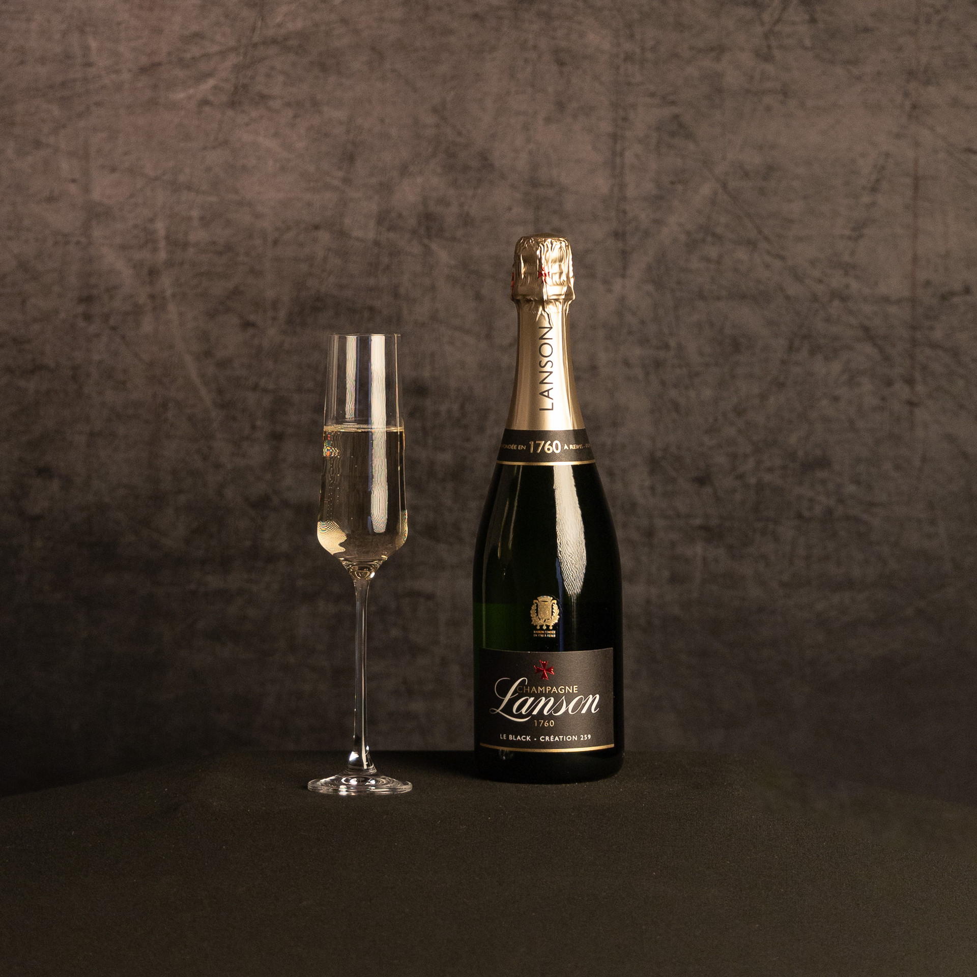 A bottle of Lanson Le Black Création 259 Champagne beside a partially filled champagne flute, photographed against a dark, textured brown backdrop.