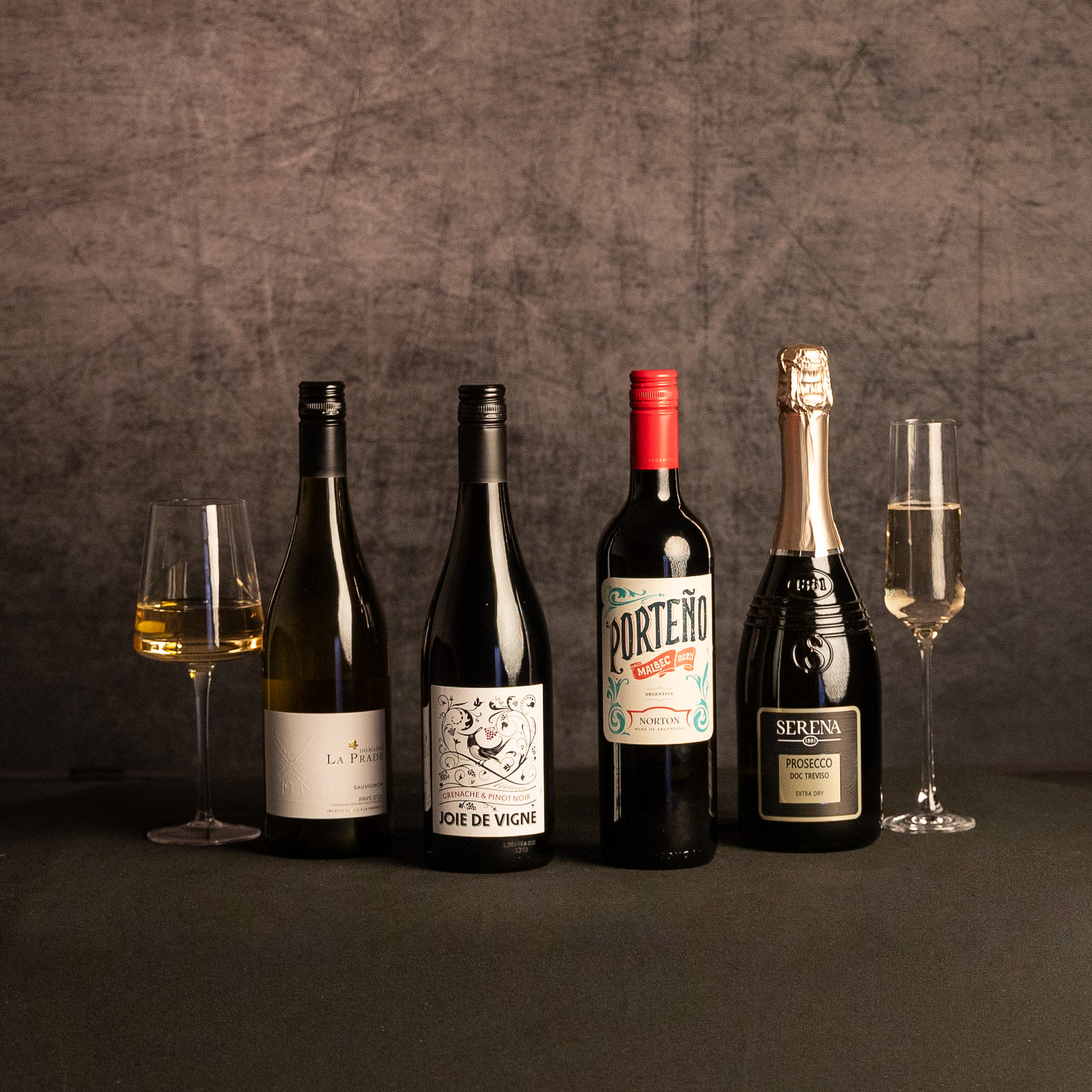 Four wine bottles—La Prade Sauvignon Blanc, Joie de Vigne Grenache & Pinot Noir, Porteño Malbec and Serena Prosecco—are arranged between glasses of white and sparkling wine against a dark, textured backdrop.