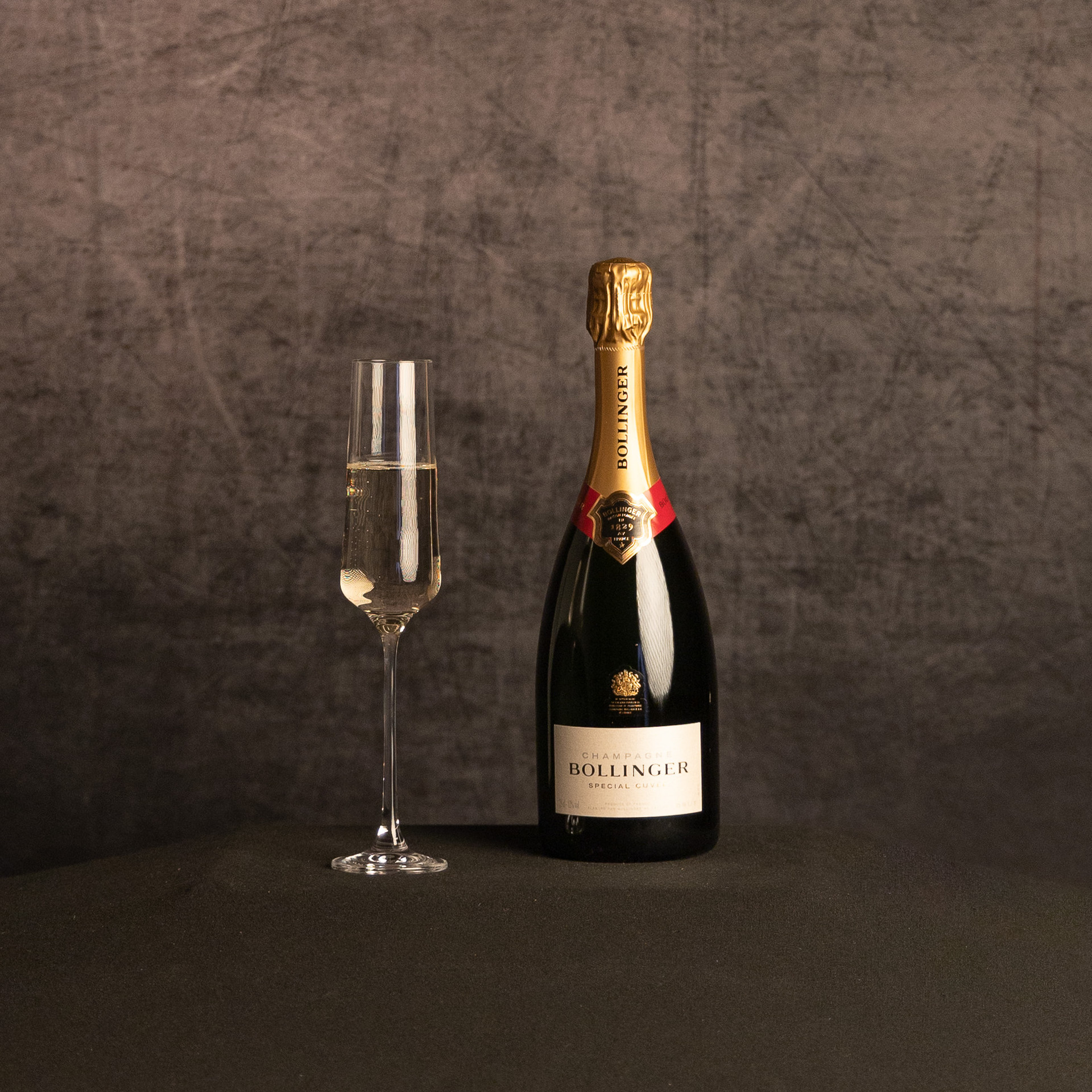 A bottle of Bollinger Special Cuvée Champagne beside a partially filled champagne flute, photographed against a dark, textured brown backdrop.