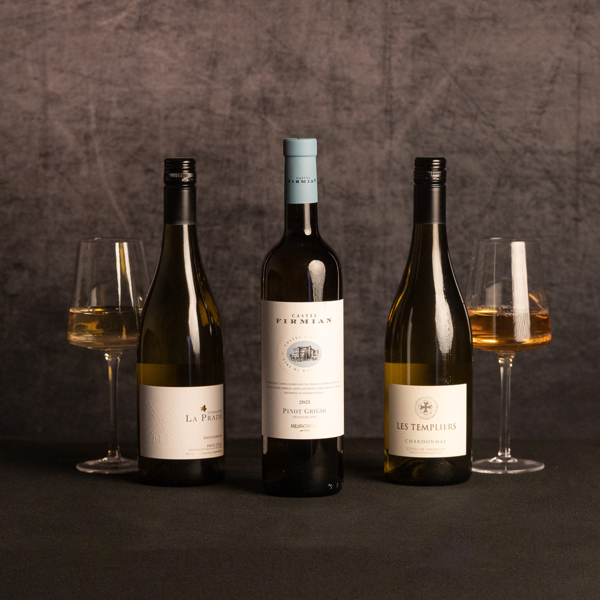Bottles of Domaine La Prade Sauvignon, Castel Firmian Pinot Grigio and Les Templiers Chardonnay are arranged between two glasses of white wine against a dark, textured backdrop.