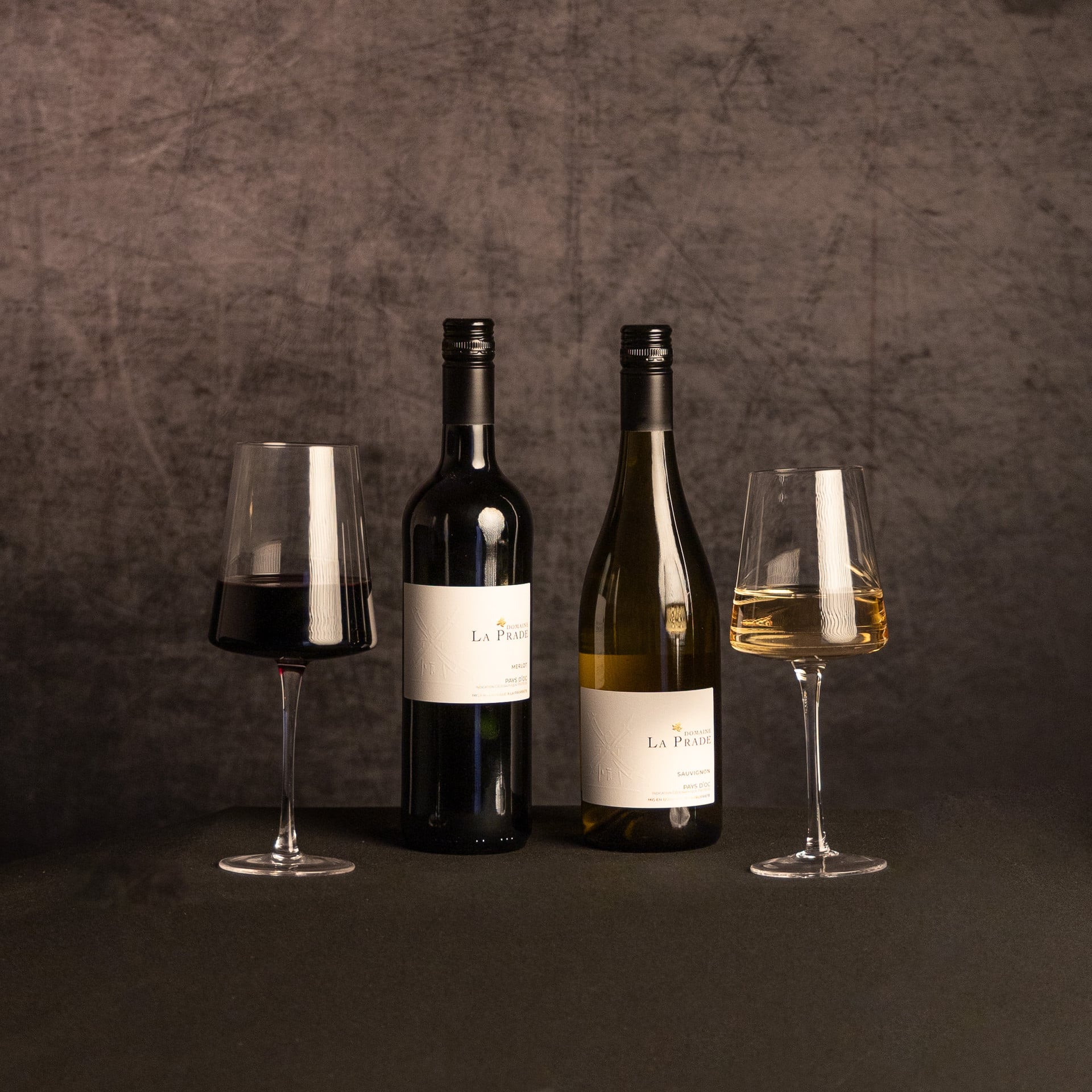 Two bottles of Domaine La Prade wine—Merlot and Sauvignon—stand between glasses of red and white wine against a dark, textured backdrop.
