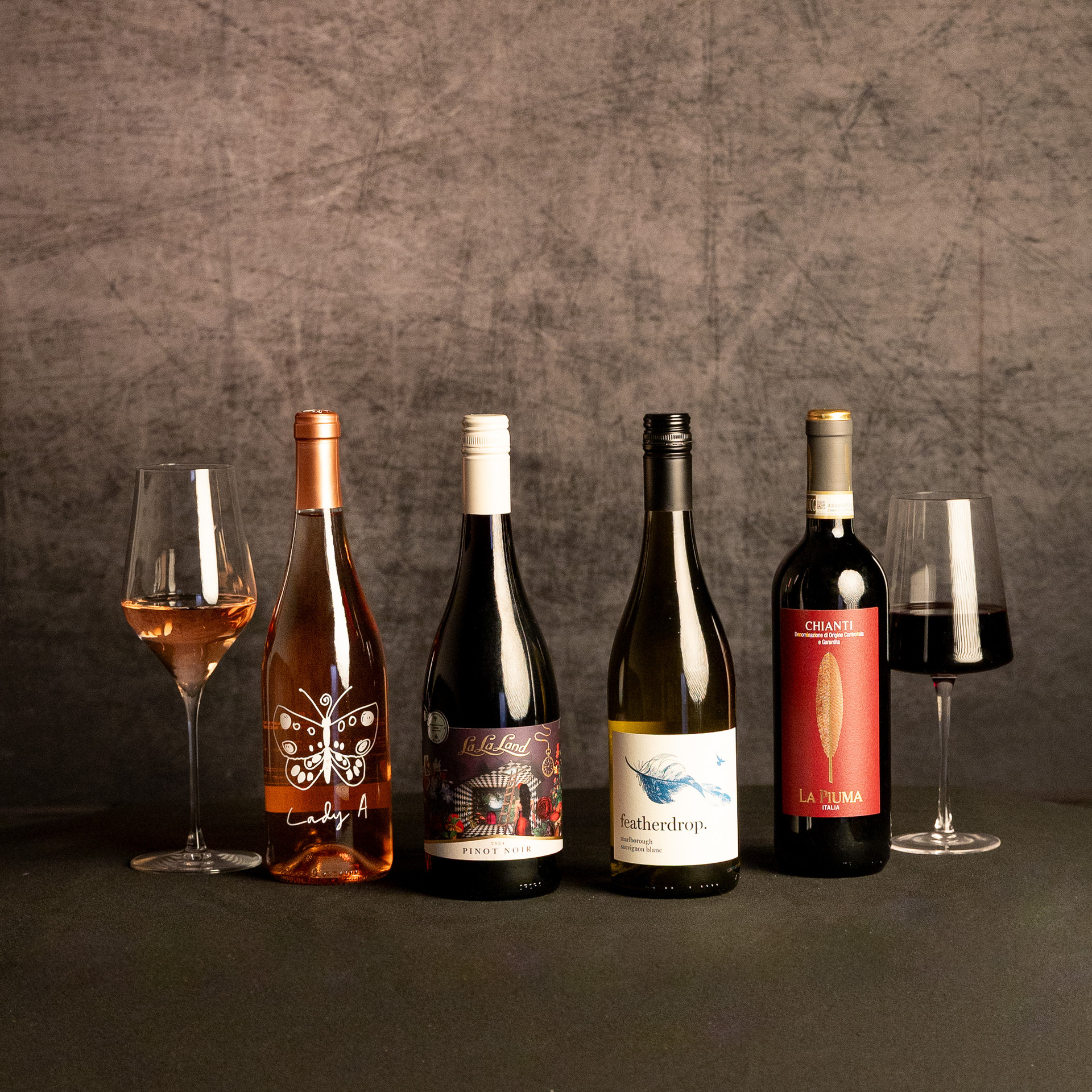 Four wine bottles—Lady A Rosé, La La Land Pinot Noir, Featherdrop Sauvignon Blanc and La Piuma Chianti—stand between glasses of rosé and red wine against a dark, textured backdrop.