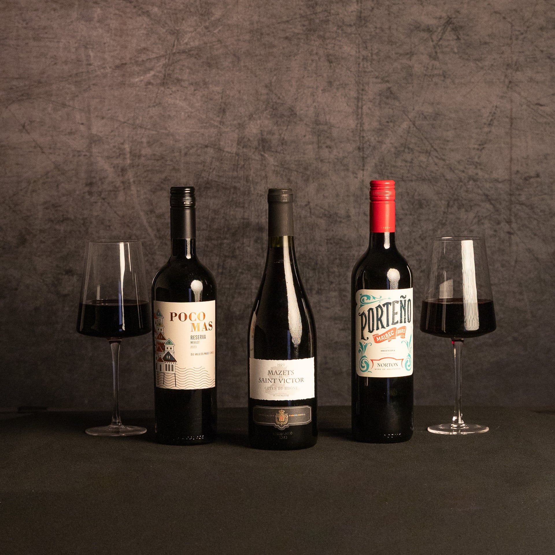 Bottles of Poco Mas Reserva Merlot, Mazets de Saint Victor Côtes du Rhône and Porteño Malbec are arranged between two glasses of red wine against a dark, textured backdrop.
