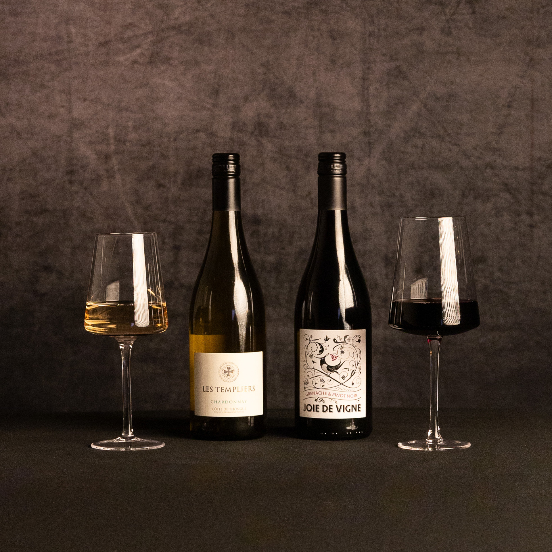Bottles of Les Templiers Chardonnay and Joie de Vigne Grenache & Pinot Noir stand between glasses of white and red wine against a dark, textured backdrop.